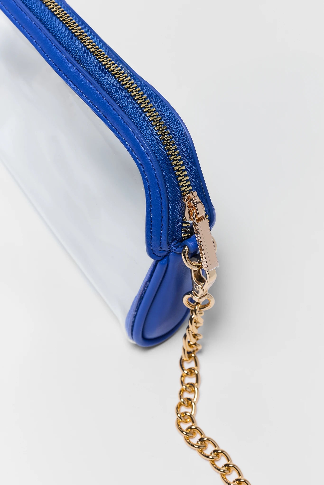 A close-up of a clear plastic pouch with a vibrant blue leather trim and a gold-tone chain strap on a white background.