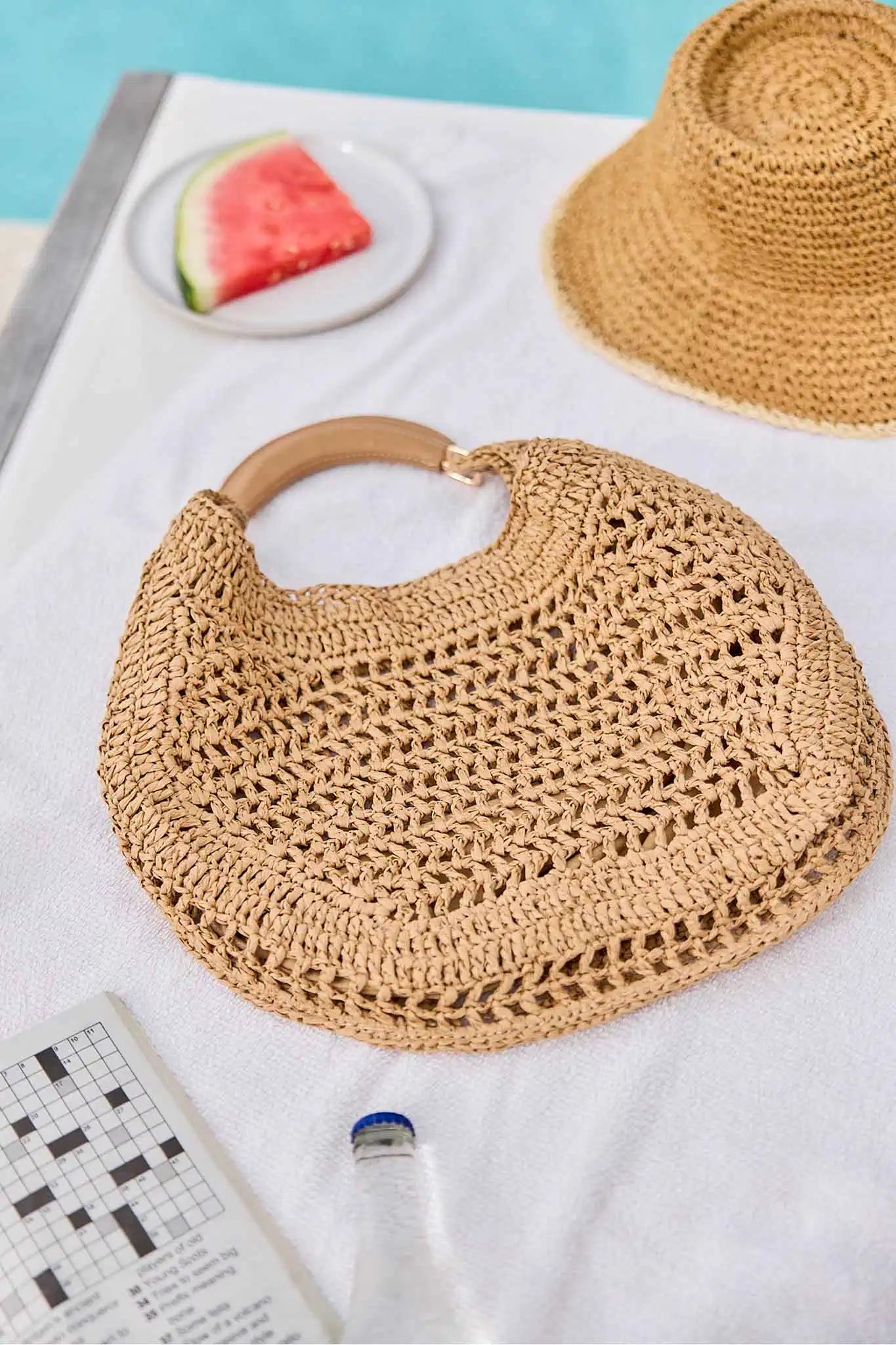 Woven handbag with wooden handle on a white surface with watermelon and hat in the background.