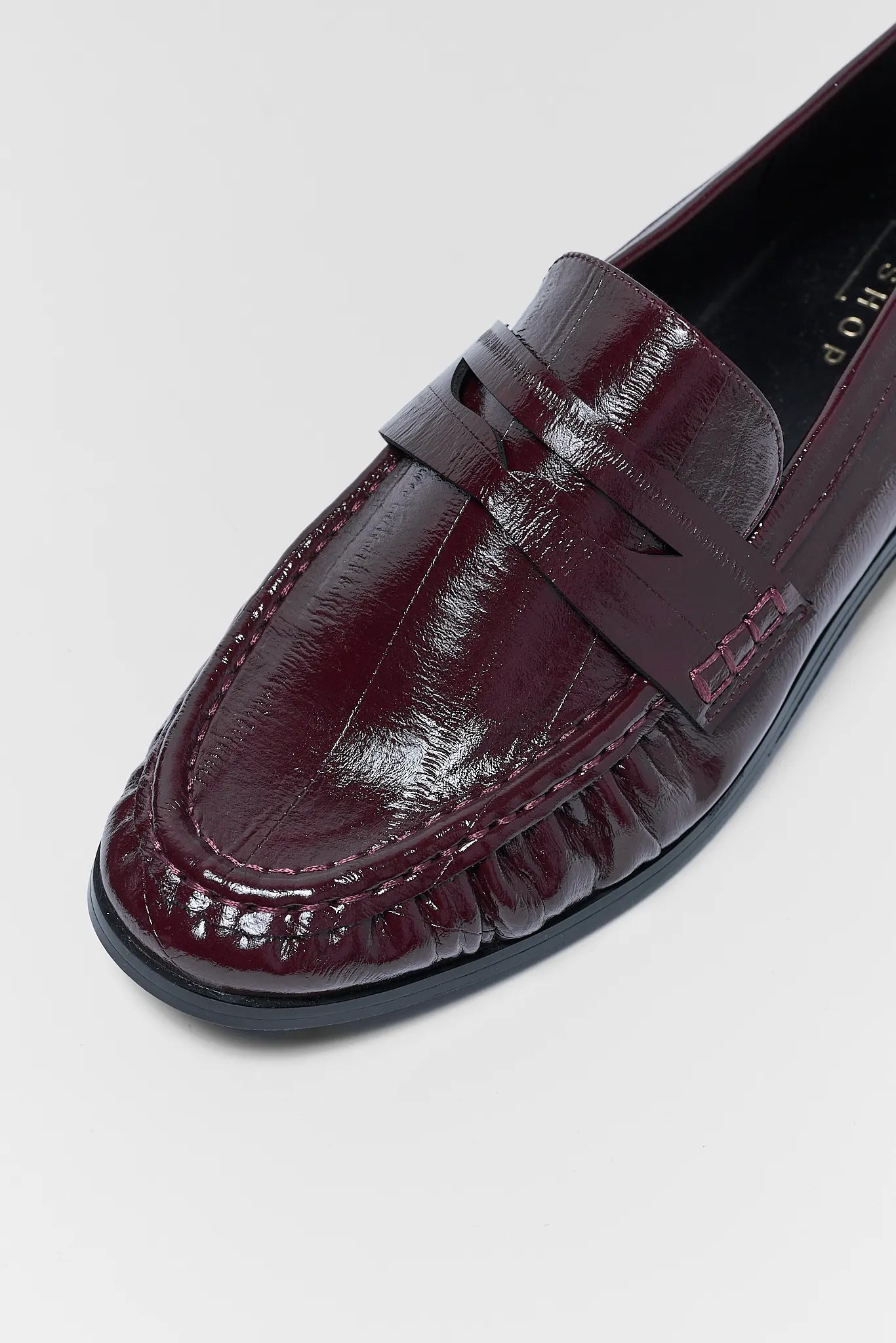 A close-up of a glossy burgundy leather penny loafer with a textured toe cap and decorative stitching, shown on a light gray background.