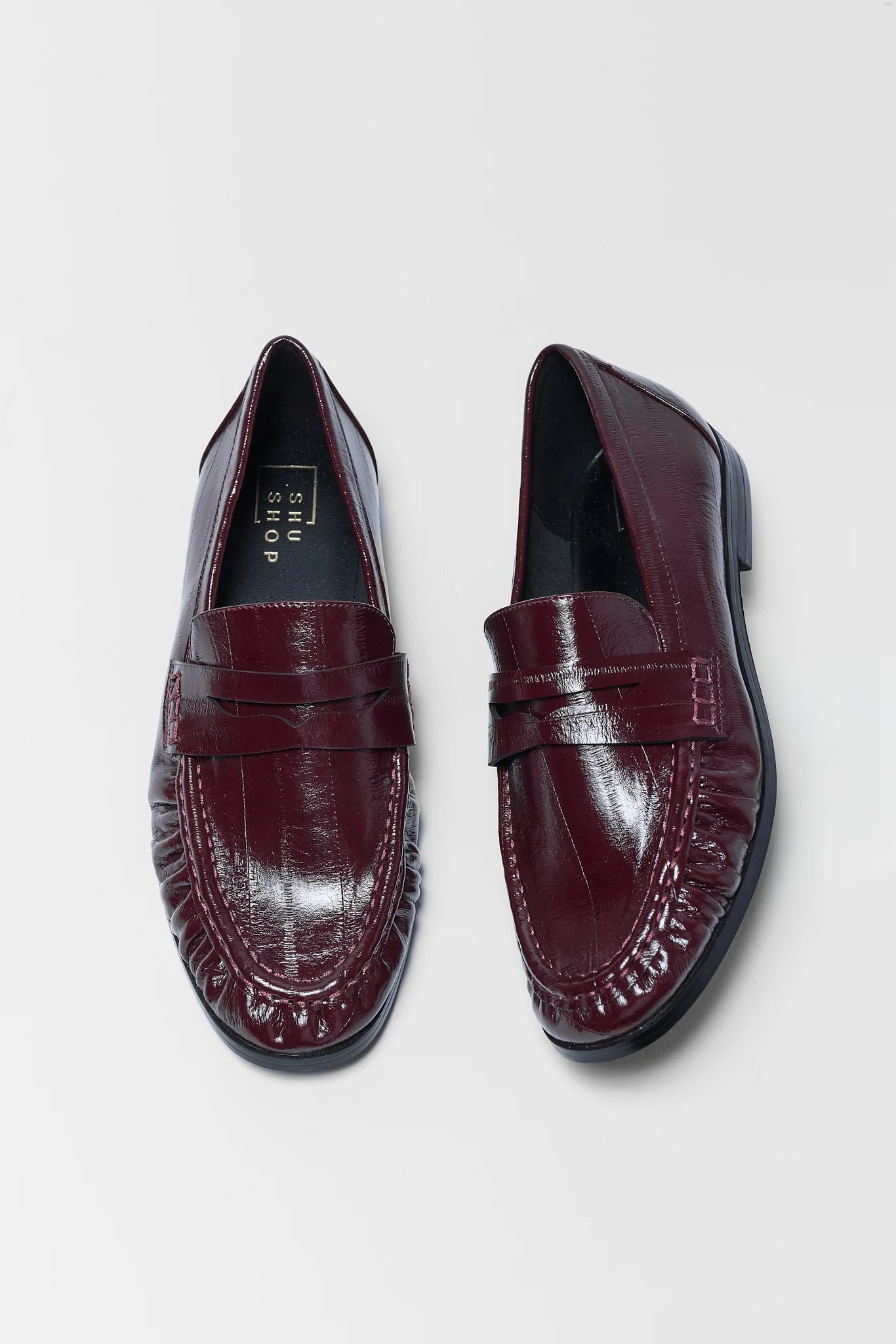 A pair of burgundy leather penny loafers with a glossy crocodile-embossed texture, shown from top and side angles on a light gray background.