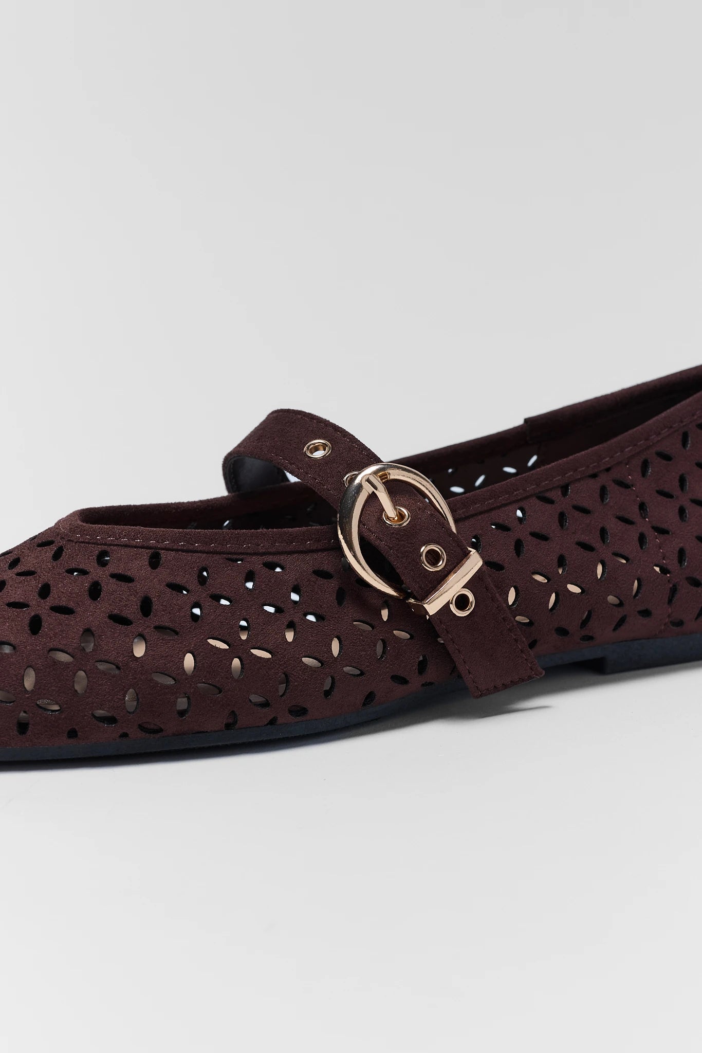 A close-up of a dark brown flat shoe with a perforated upper and a gold-tone buckle strap, shown against a light gray background.