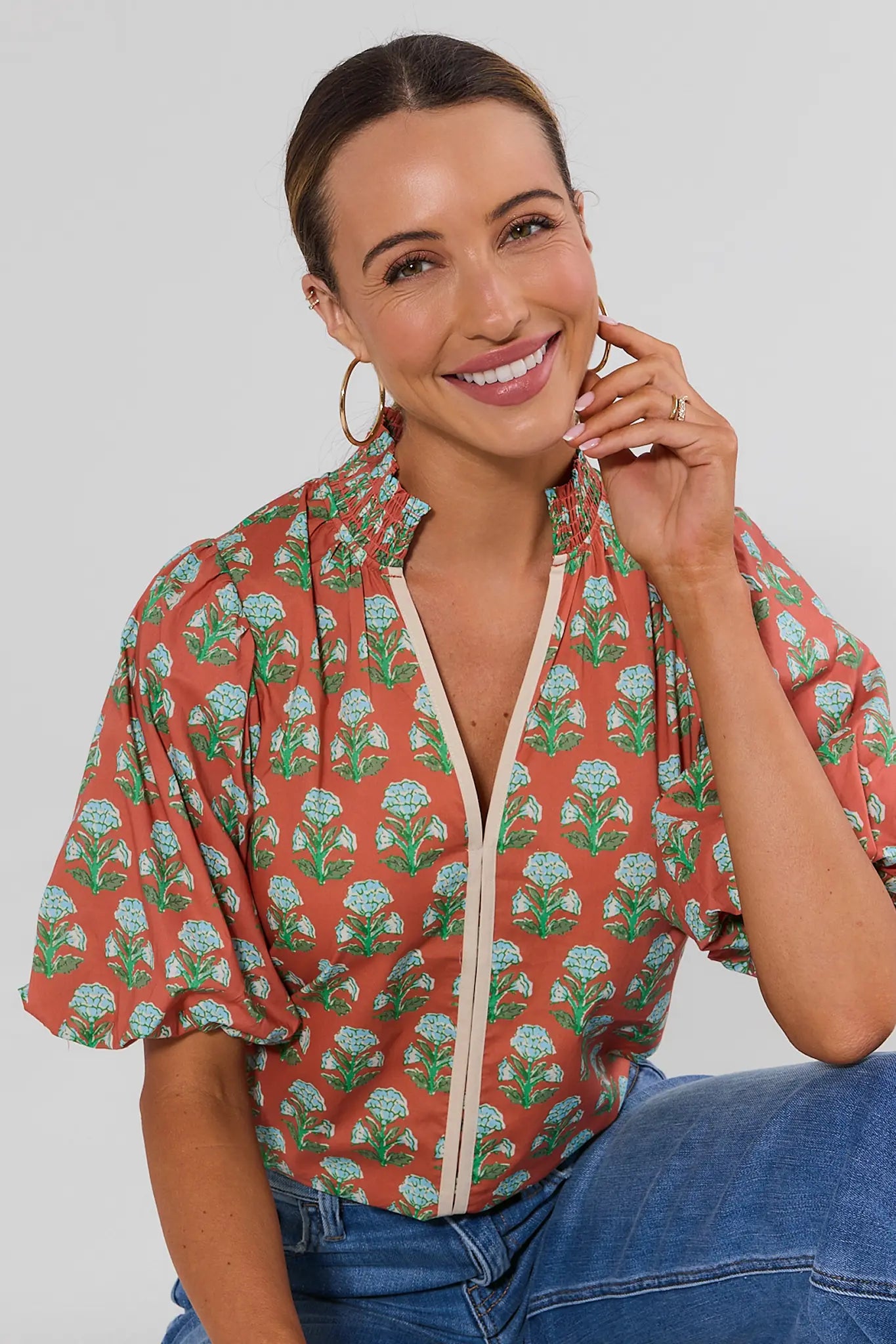 A woman sitting wearing a short-sleeve button-up blouse with a terracotta orange base, green leafy floral pattern, and light blue flower details, paired with blue jeans against a light gray studio background.