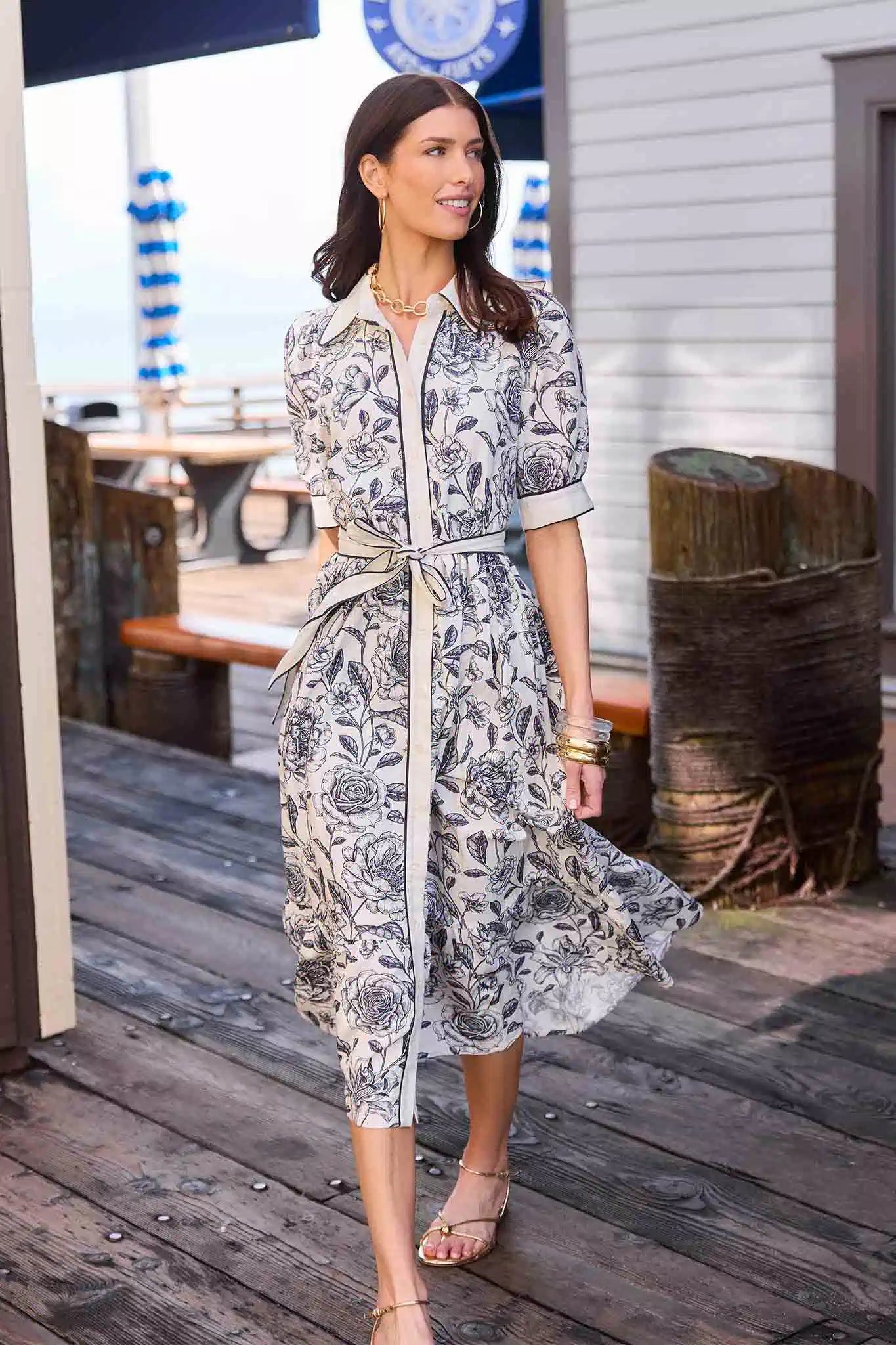Woman in a floral dress standing on a wooden deck with a coastal background.