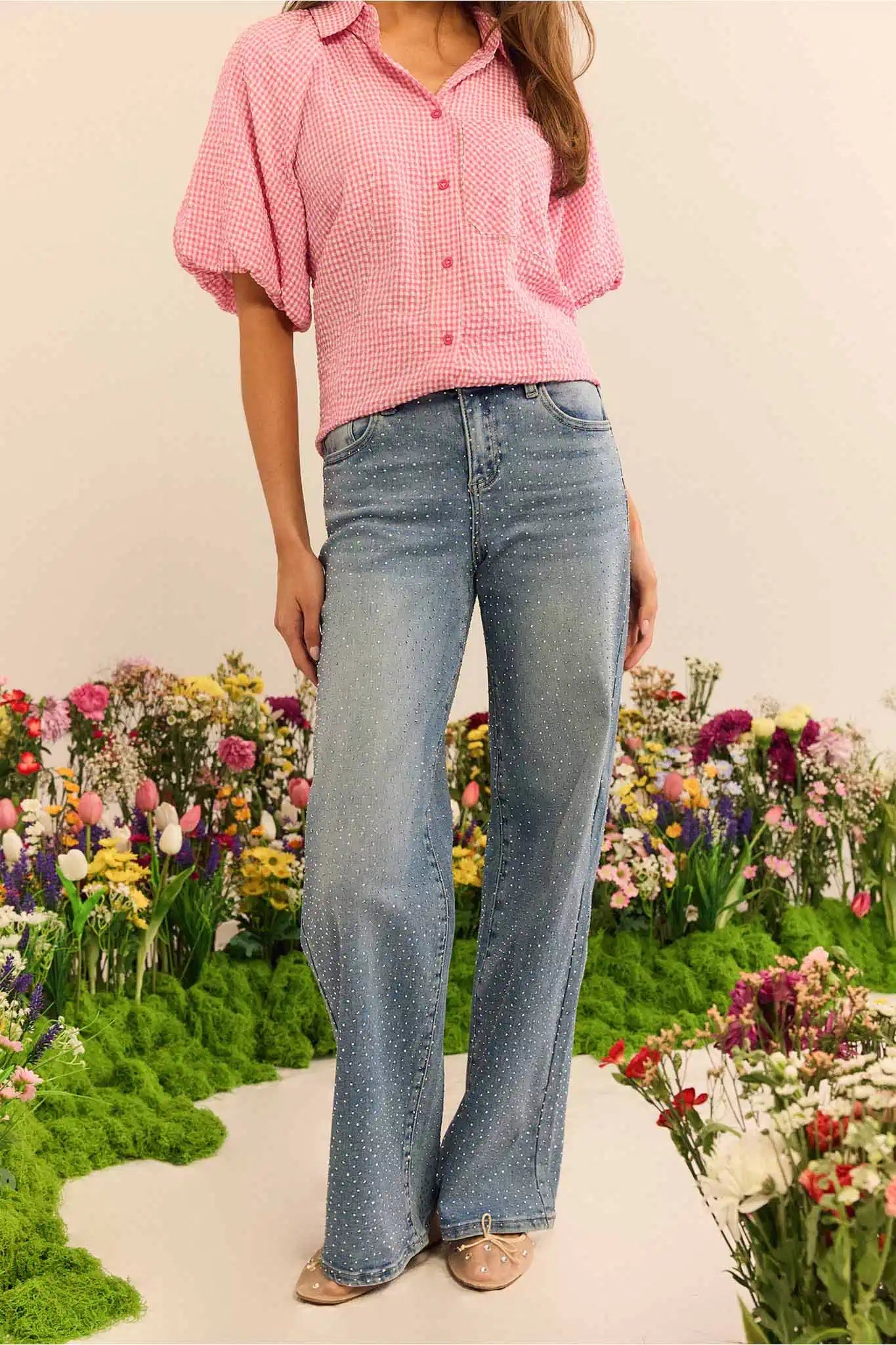 Person wearing a pink checkered shirt and blue jeans standing in front of a floral garden.