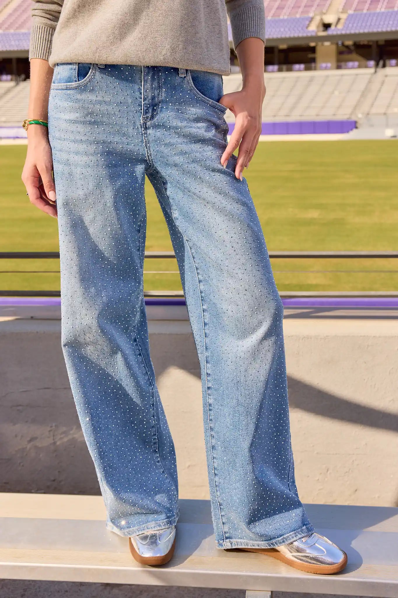 A person wearing light-wash wide-leg denim jeans with a speckled texture, paired with a beige sweater and metallic silver shoes, standing on a concrete structure in an outdoor stadium setting.