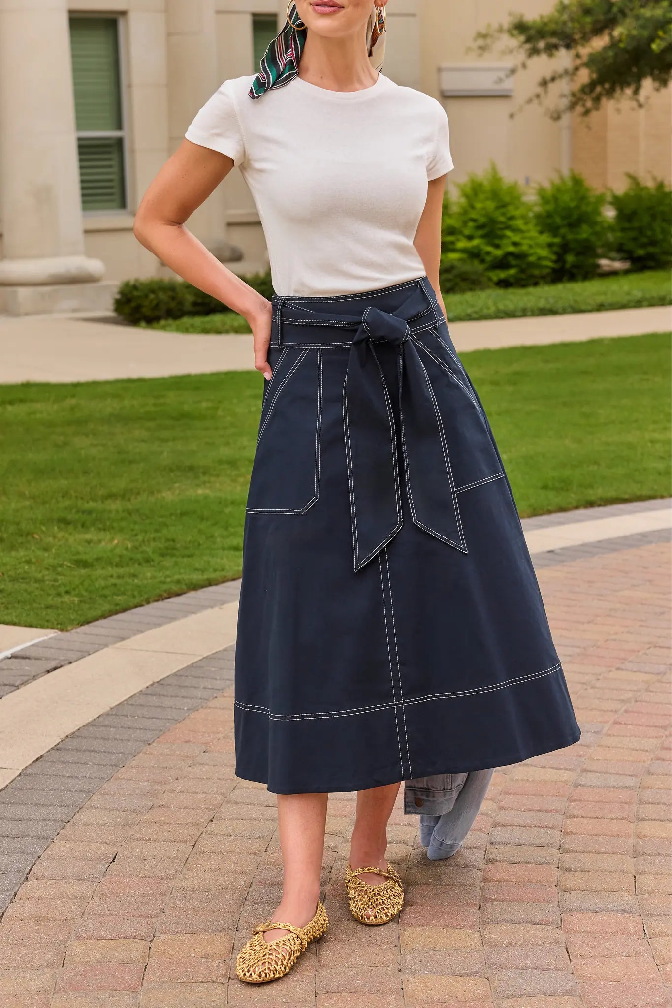 A woman wearing a white short-sleeve t-shirt and a high-waisted navy blue midi skirt with white contrast stitching and a side tie, paired with leopard-print flat shoes, standing on a brick pathway outdoors.