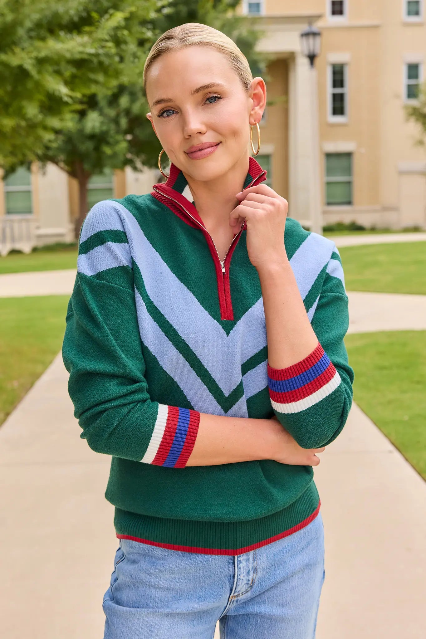A woman wearing a green quarter-zip sweater with a large light blue chevron pattern and red, white, and blue striped cuffs, standing outdoors on a campus walkway.