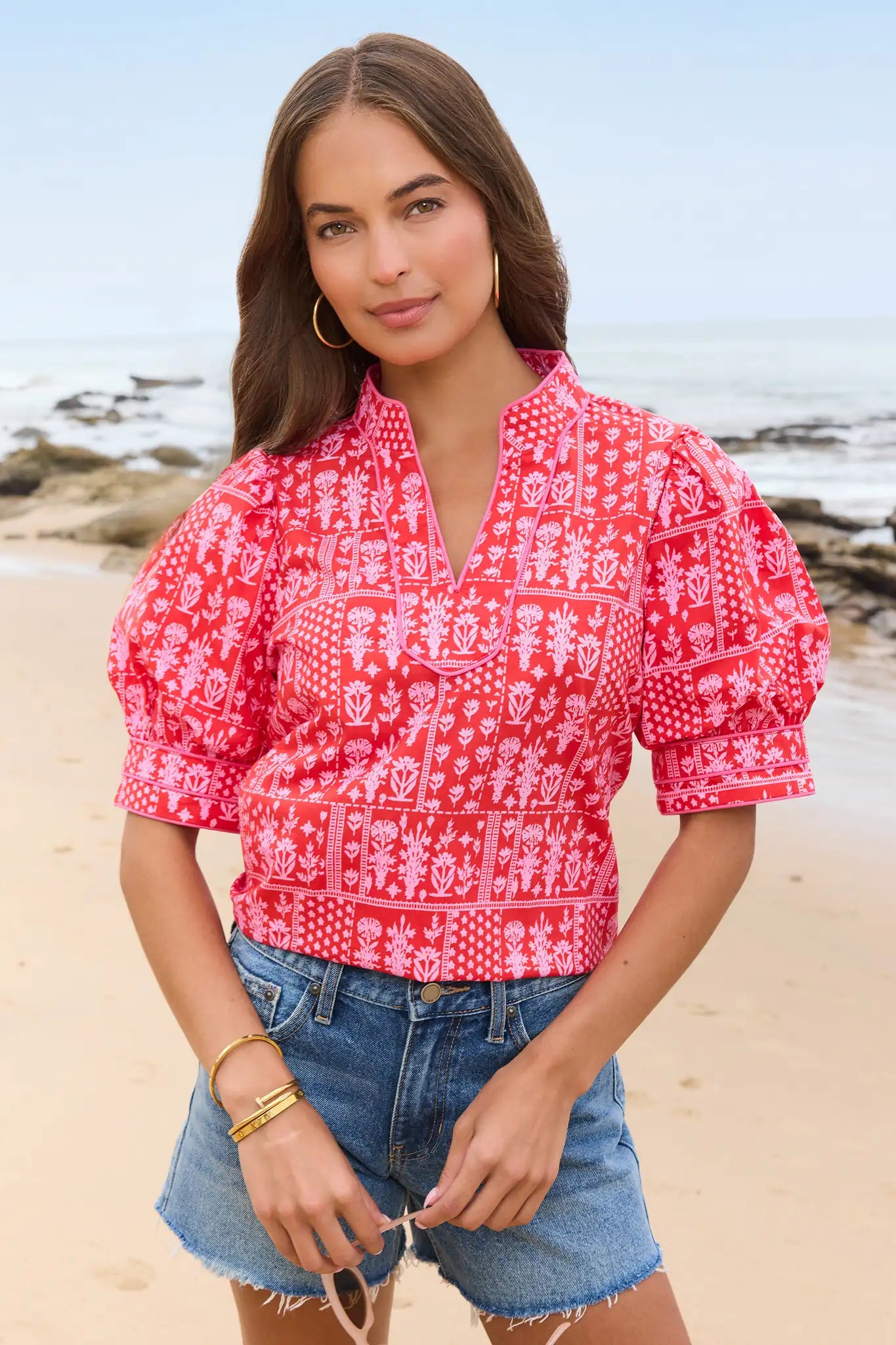 Woman wearing a red patterned blouse and denim shorts on a beach.