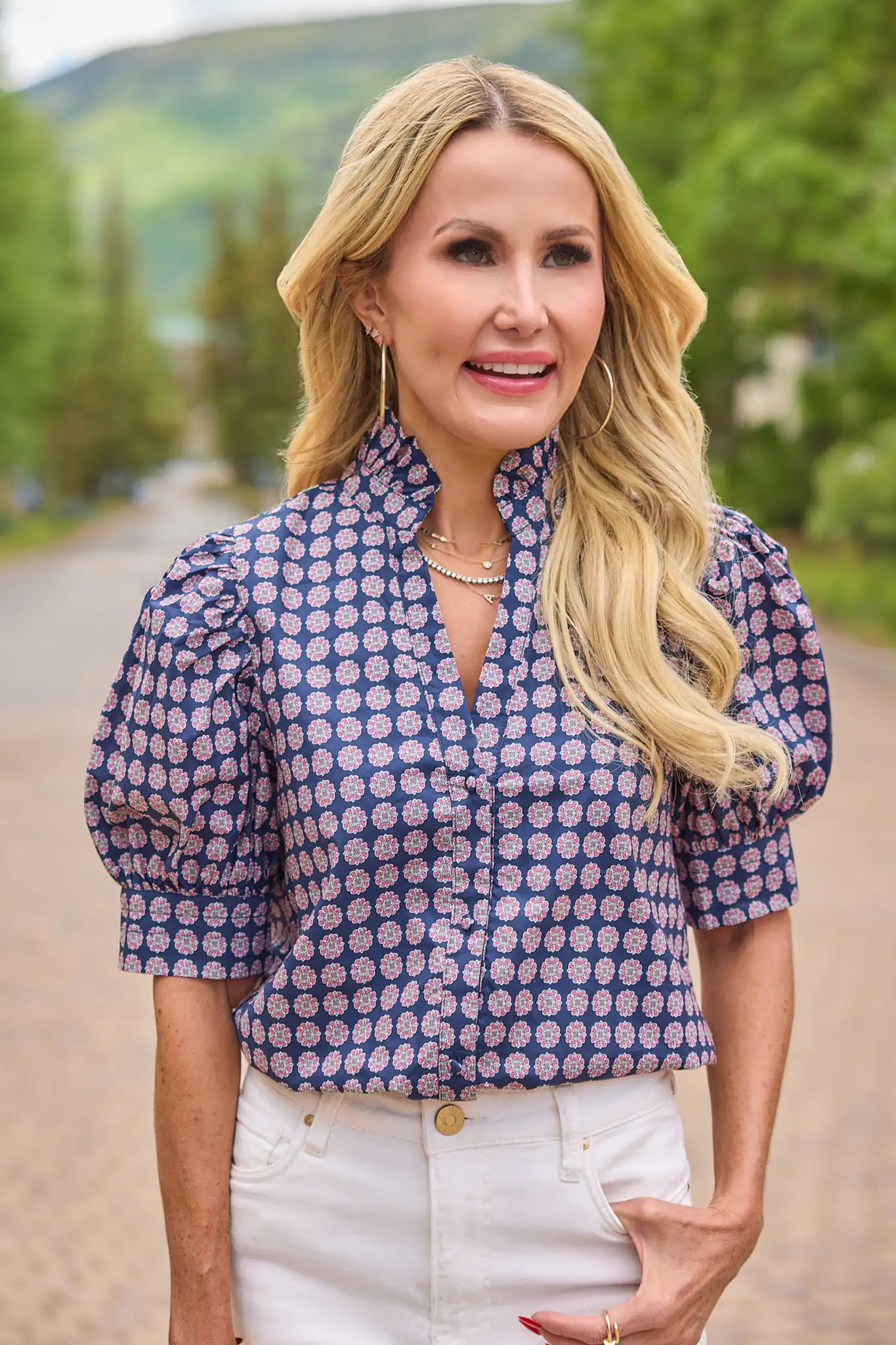 A woman with blonde hair wearing a blue and white patterned puff-sleeve blouse and white pants, standing outdoors on a path with trees and hills in the background.