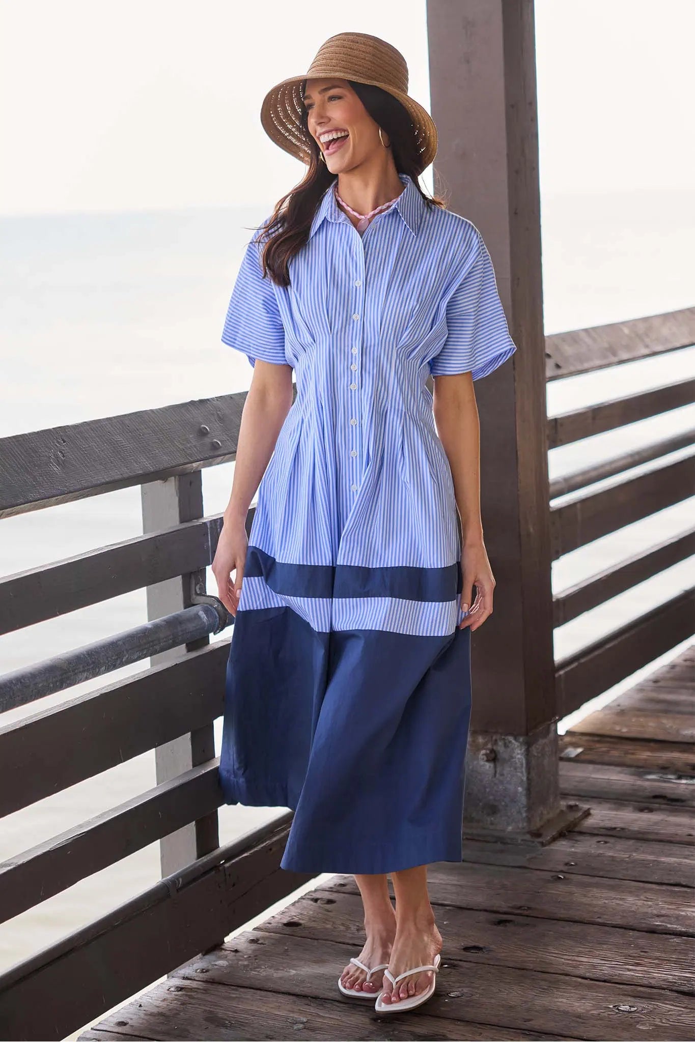 Woman in a blue striped shirt and wide-brimmed hat standing on a wooden deck.