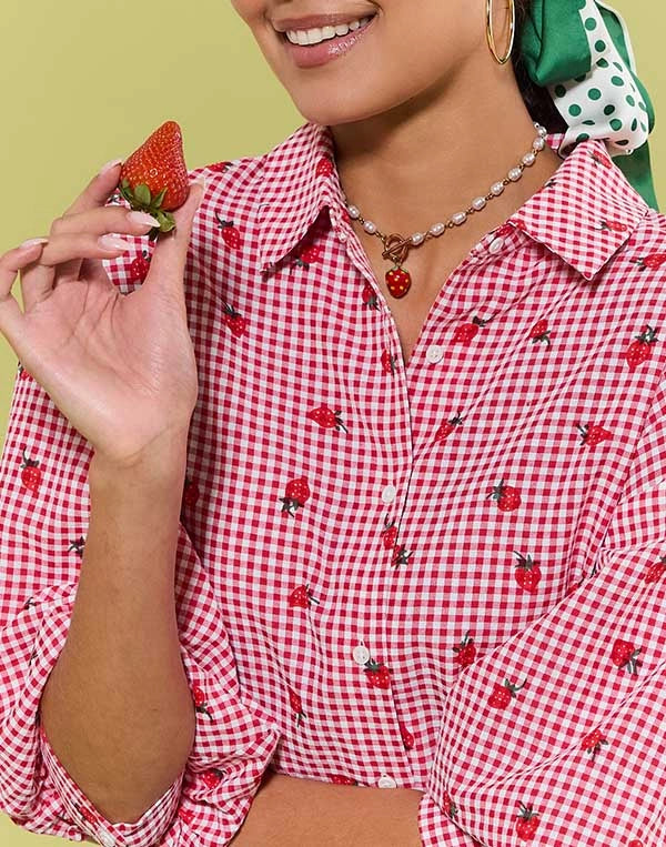 Person wearing a red and white checkered shirt with strawberry pattern, holding a strawberry.