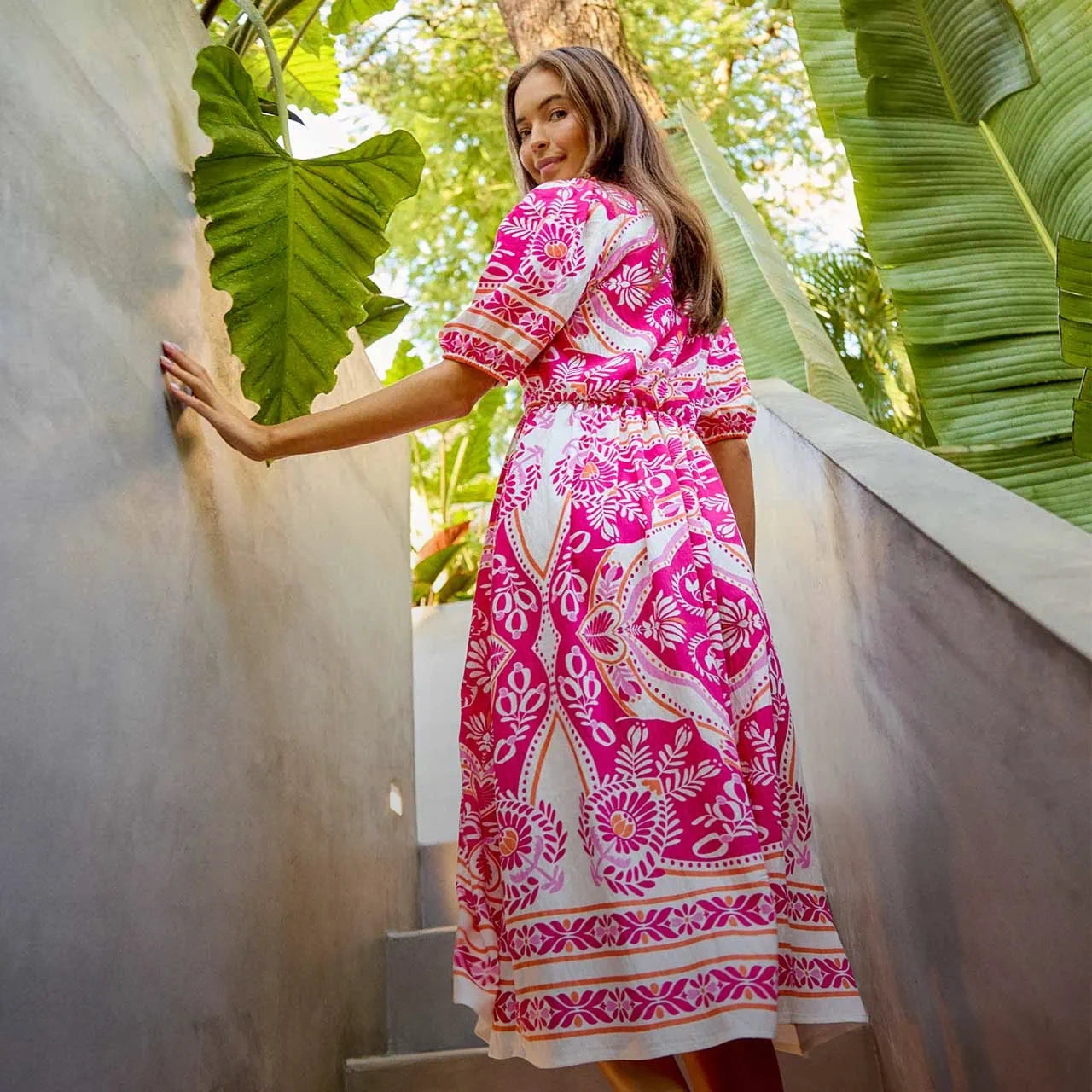 Woman in a pink and white patterned dress standing outdoors with greenery.