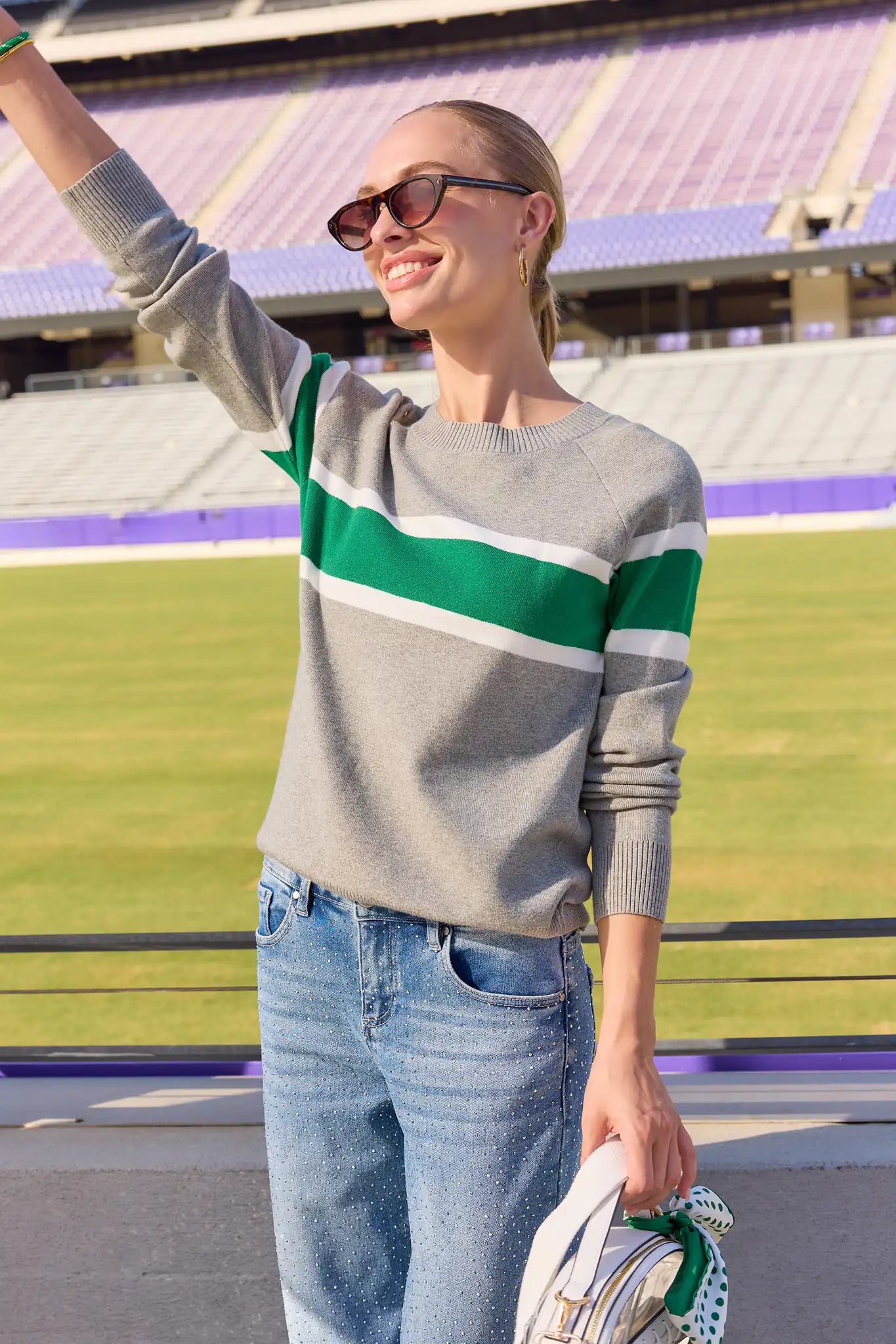 A woman stands on a sports field wearing a gray knit sweater with green and white horizontal stripes, blue jeans, and sunglasses, holding a handbag.