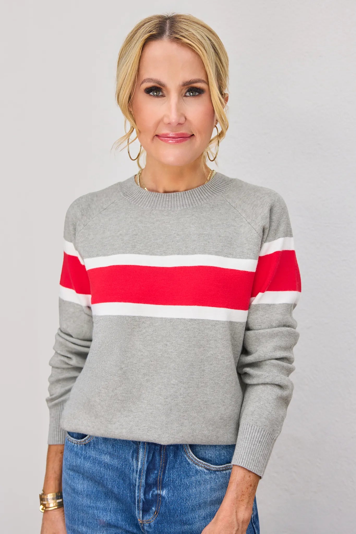 A woman wearing a heather gray knit sweater with a bold horizontal red and white stripe across the chest, paired with blue jeans, against a neutral light gray background.