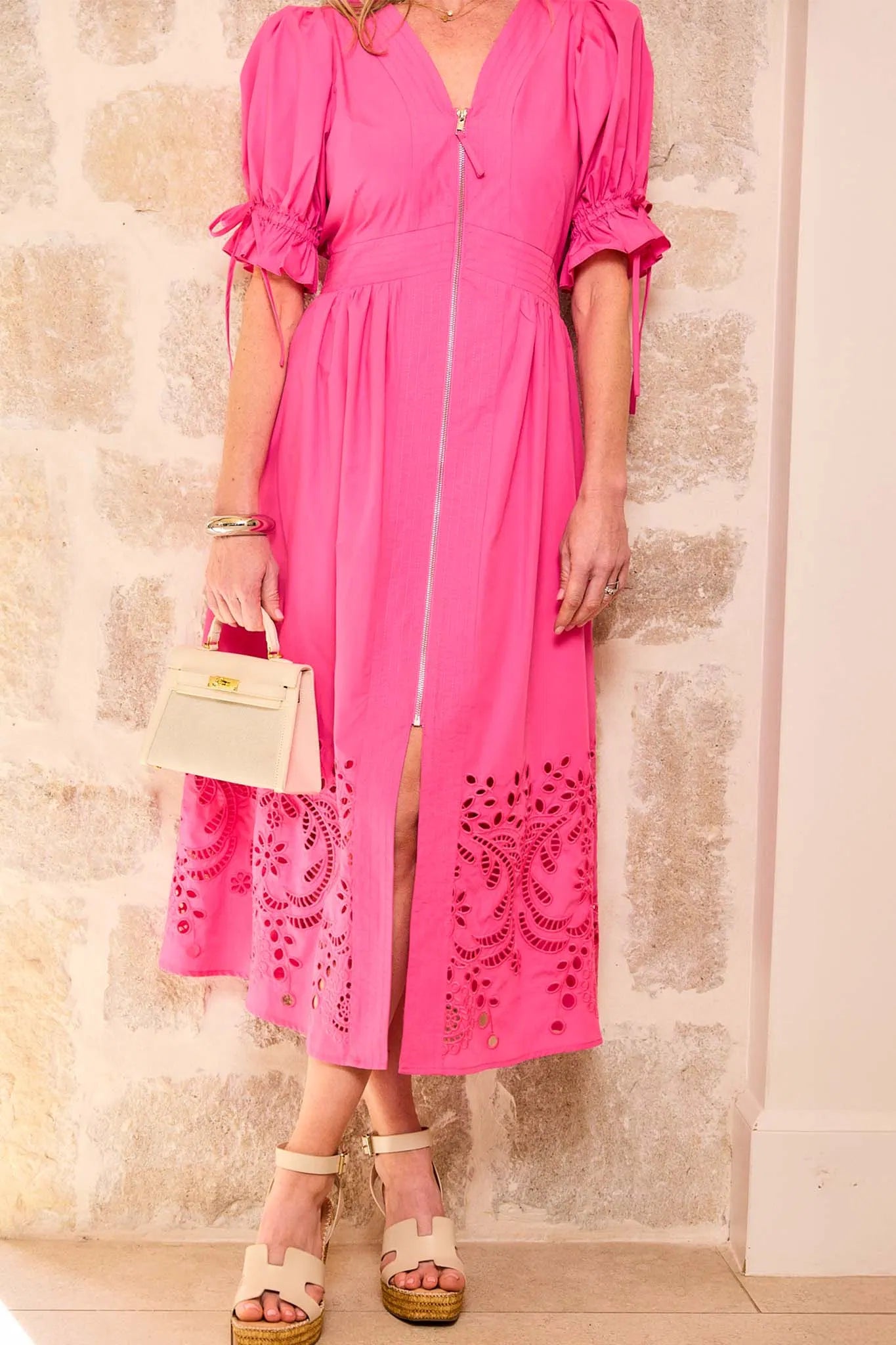 Person wearing a pink dress with intricate cut-out patterns, holding a small handbag against a stone wall.