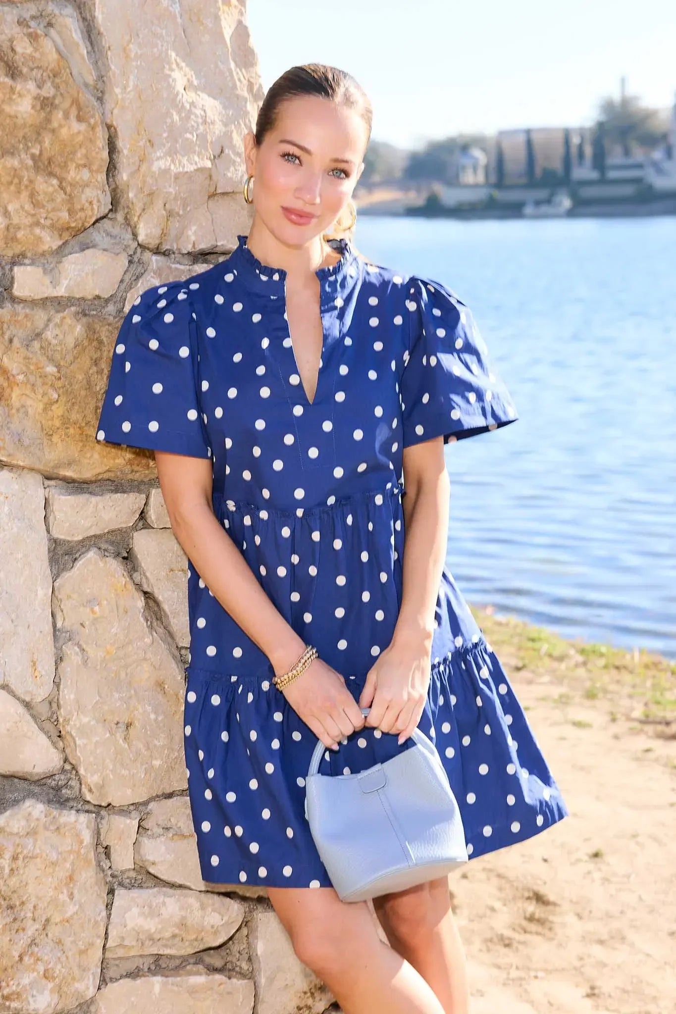 Woman in a blue polka dot dress standing by a stone wall with water in the background