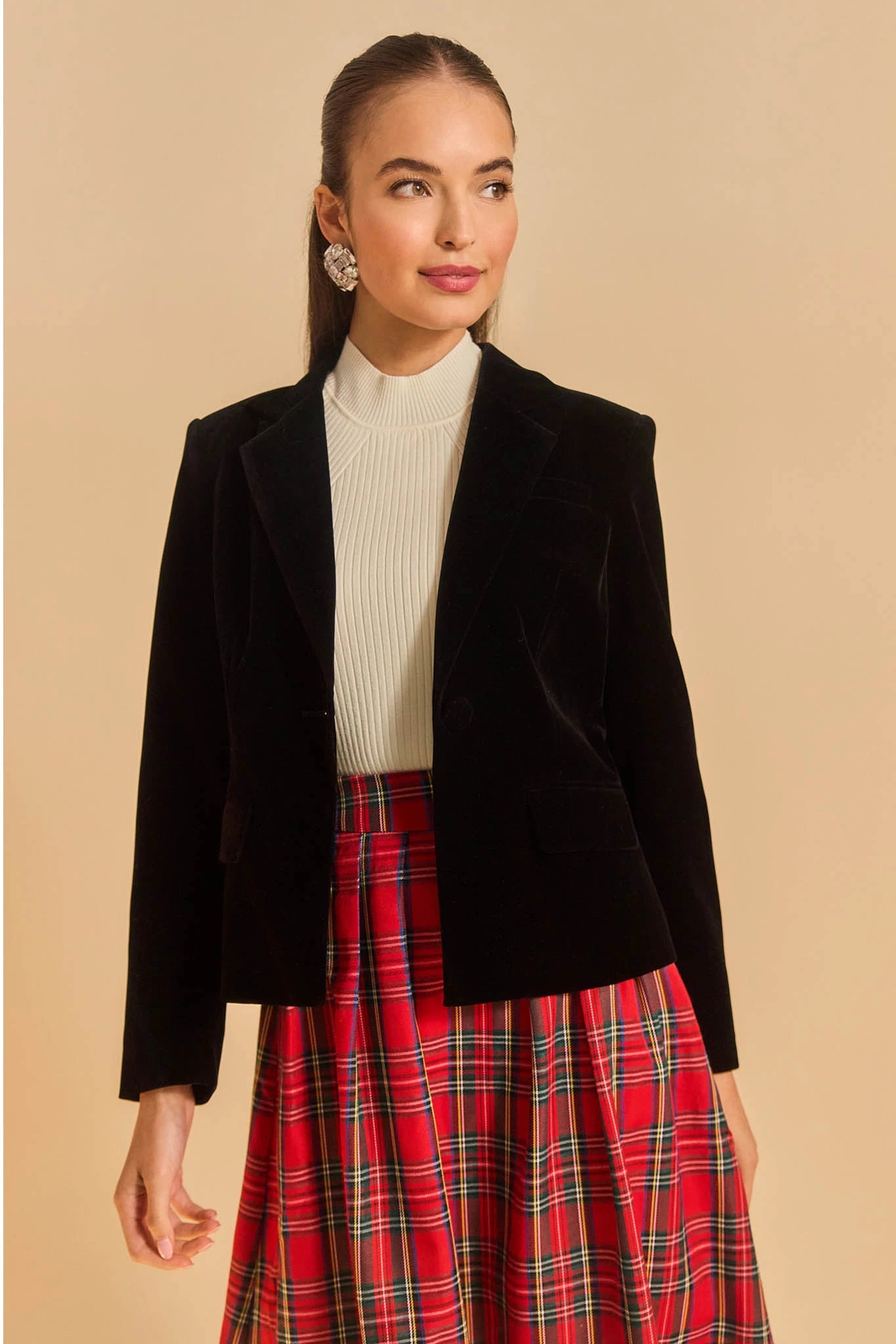 Woman wearing a black blazer, white turtleneck, and red plaid skirt on a beige background