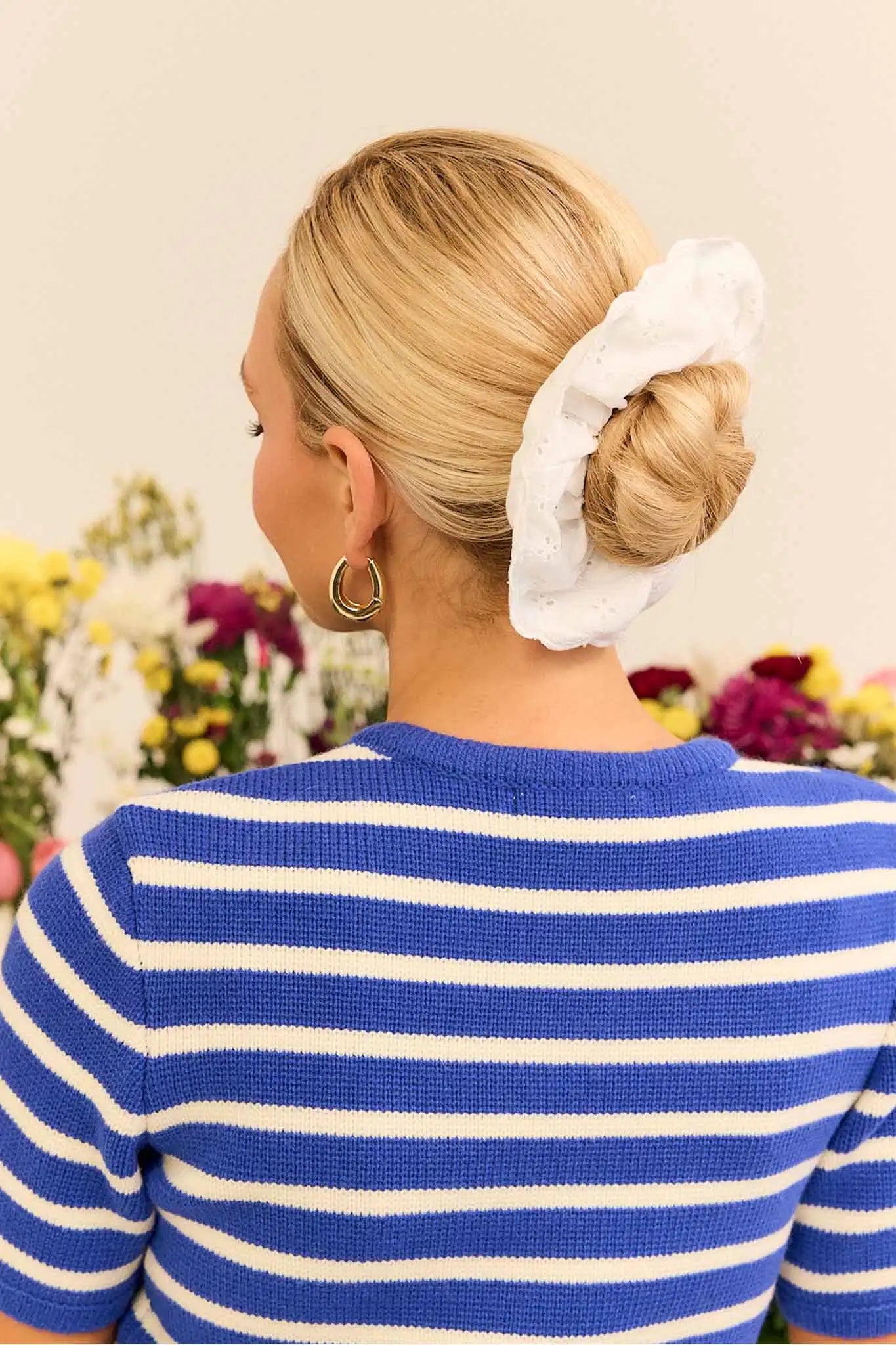 Woman with a bun wearing a white scrunchie, standing in front of flowers.