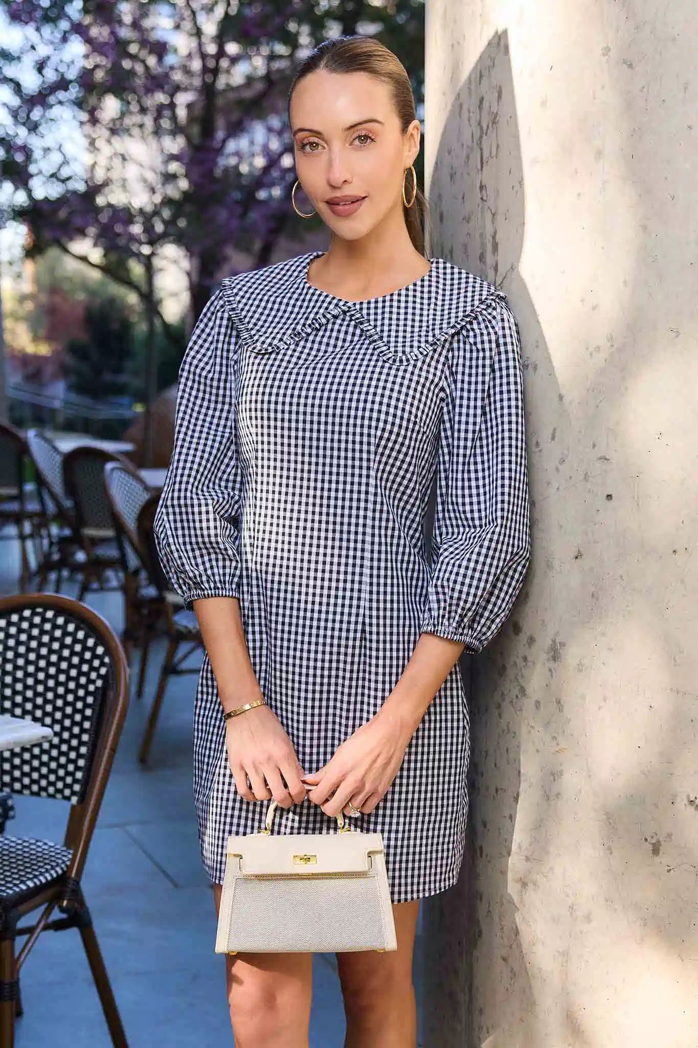 Woman in a blue checkered dress holding a white handbag against a stone wall.