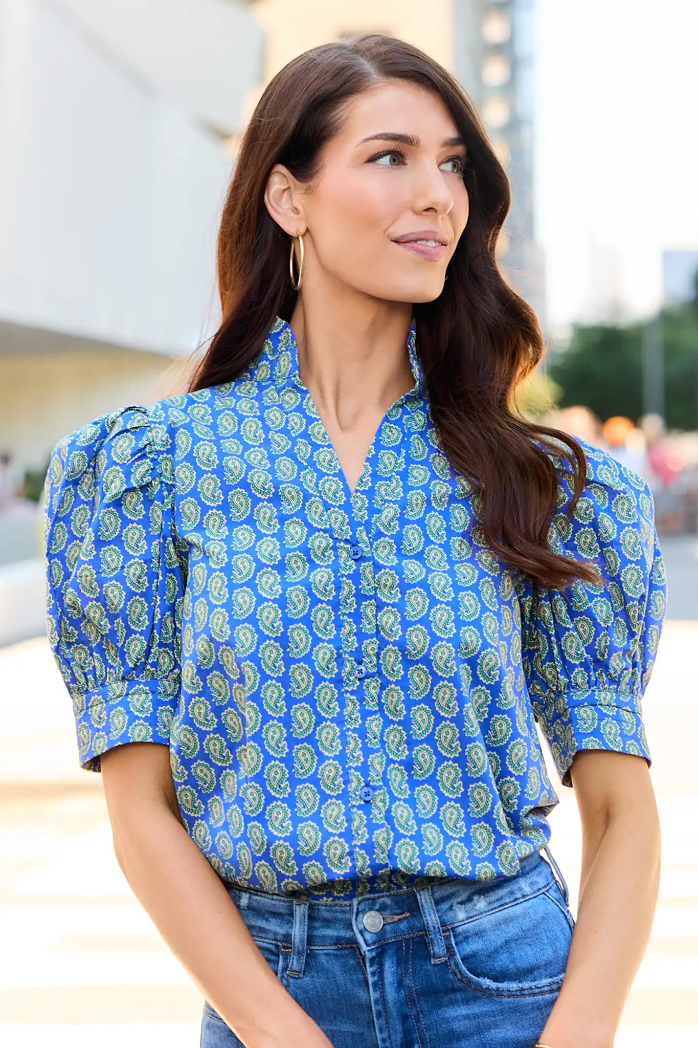 A woman wearing a blue short-sleeve button-up blouse with a green and white paisley pattern and puff sleeves, paired with blue jeans.