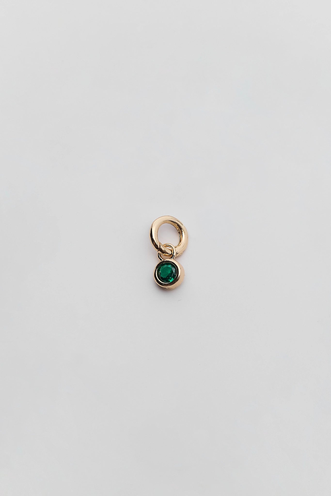 A small gold charm featuring a round green gemstone in a bezel setting, suspended from a simple gold ring, shown on a plain light gray background.