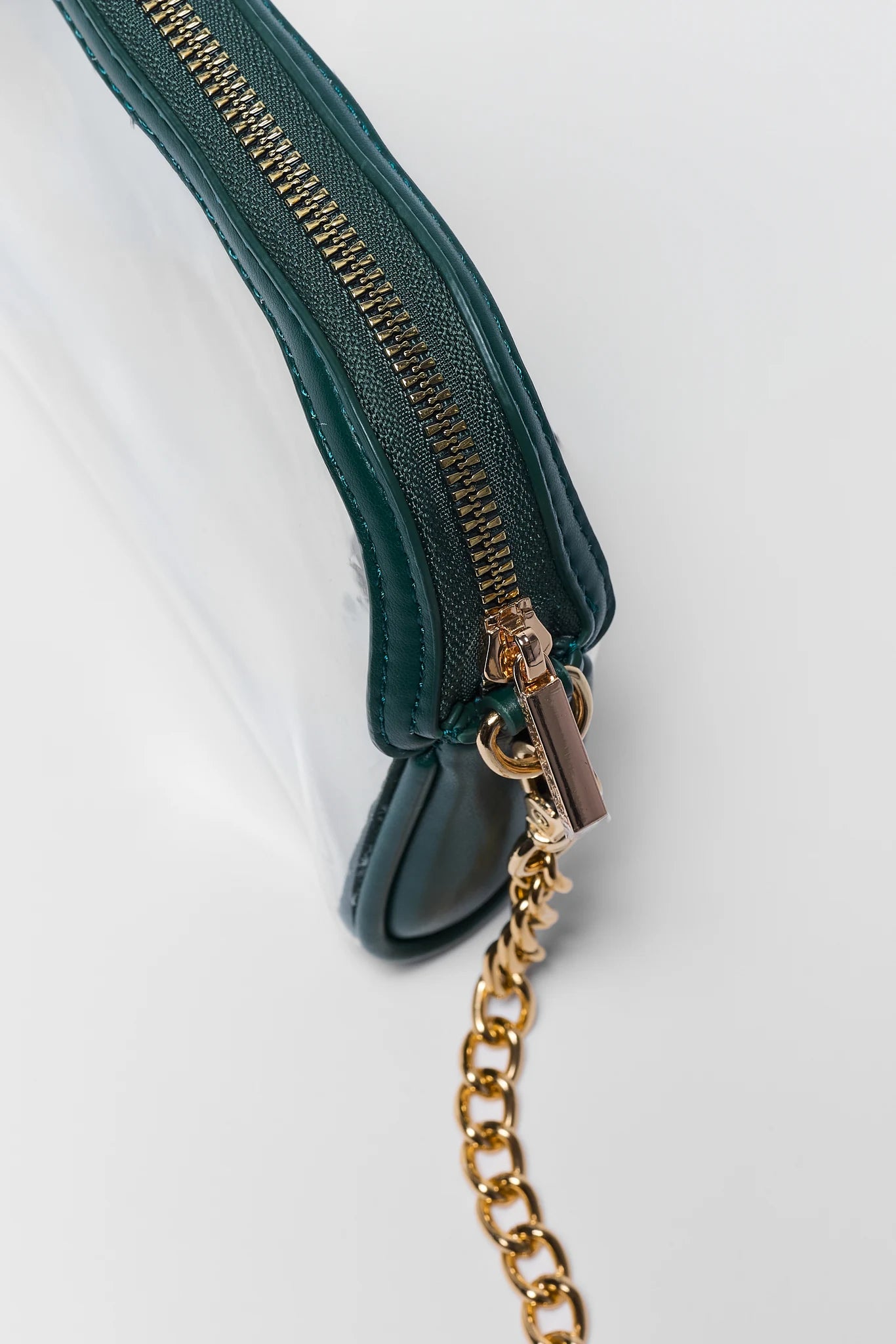 Close-up of a dark green leather zippered pouch with a gold-tone chain strap and a clear plastic body, shown on a light gray background.