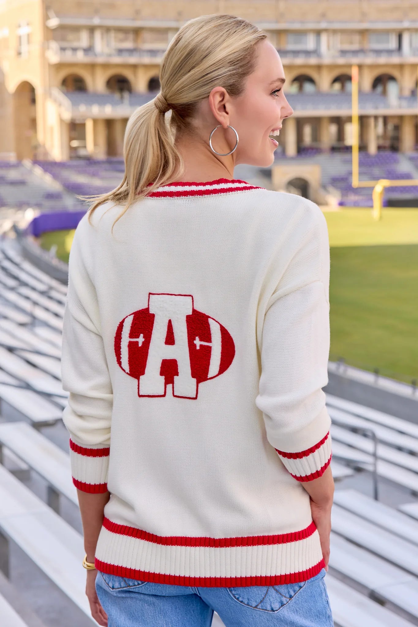 A woman wearing a cream knit sweater with red and white athletic trim and a large red and white football-shaped 'A' graphic on the back, standing in stadium seating.