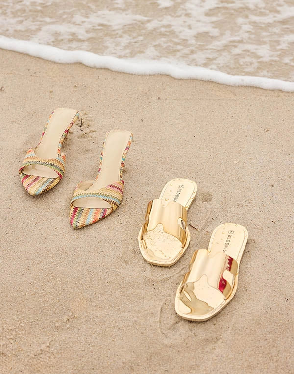 Pair of colorful sandals and a gold sandal on a sandy beach with ocean waves.