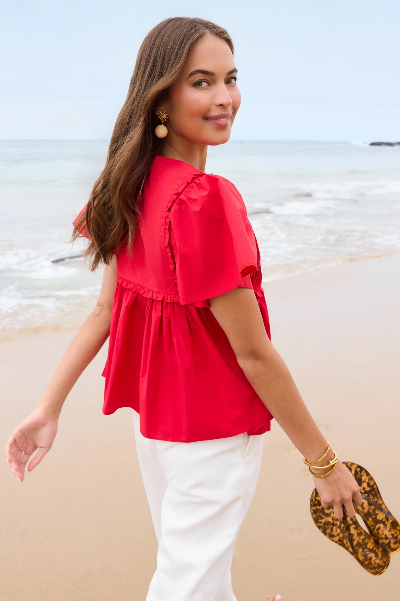Woman in a red blouse and white pants standing on a beach.