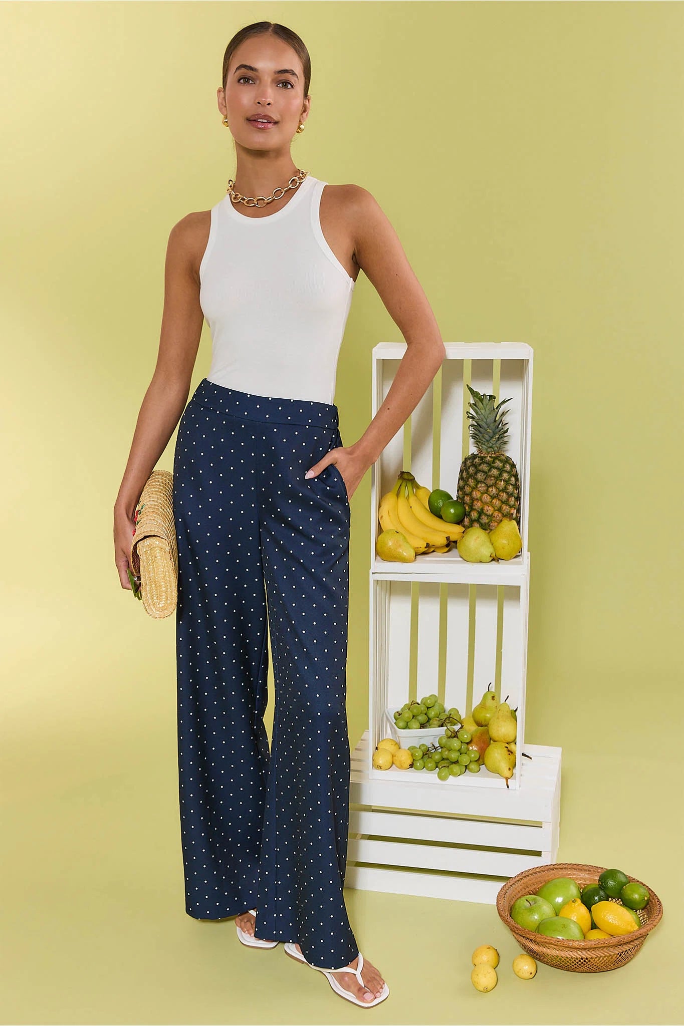 Woman in a white sleeveless top and navy polka dot pants standing against a light green background with a fruit display.