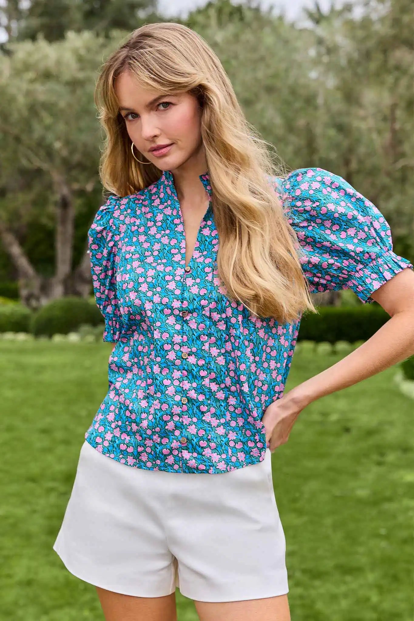 Woman wearing a floral blouse and white shorts standing outdoors with greenery in the background