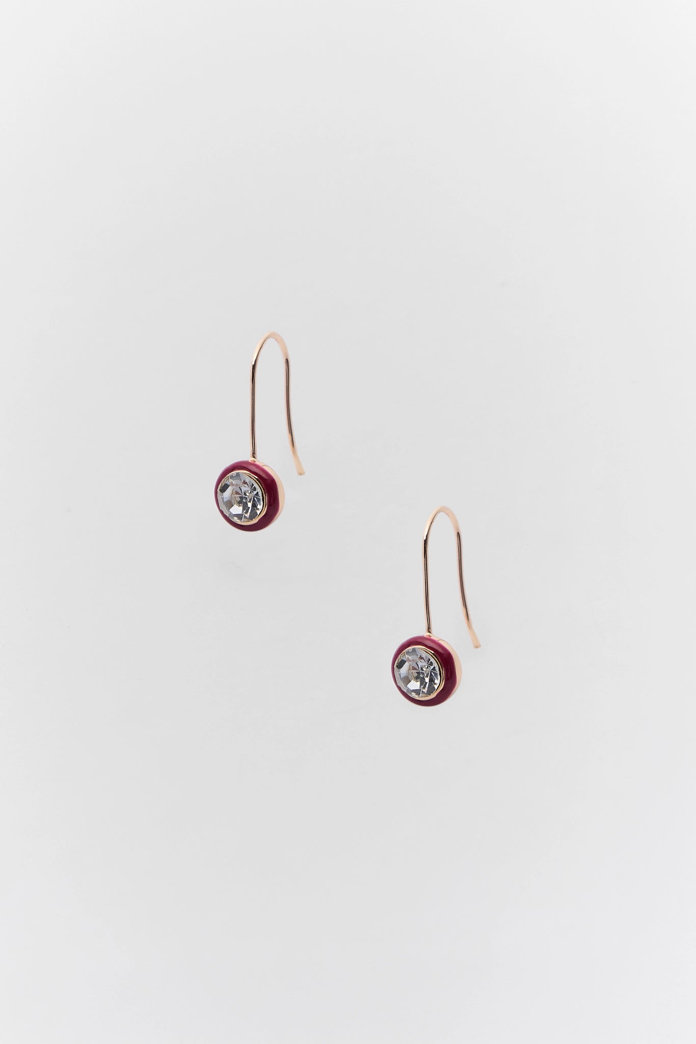 A pair of rose gold drop earrings featuring a small circular red enamel bezel with a clear faceted gemstone, suspended from thin rose gold hooks against a light gray background.