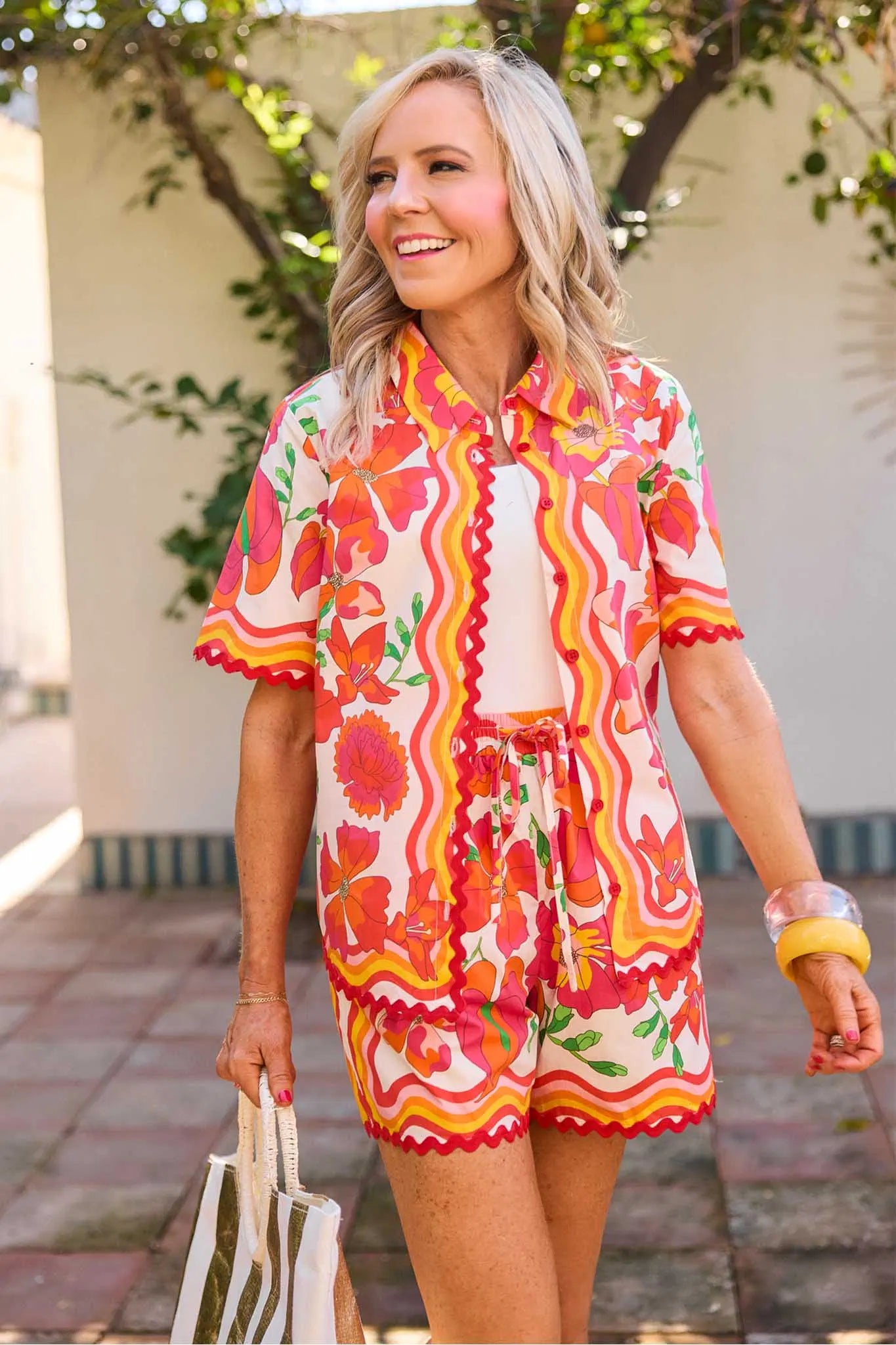 Woman wearing a colorful Melissa Orange Printed Button Up Top
and shorts with a patterned bag outdoors.