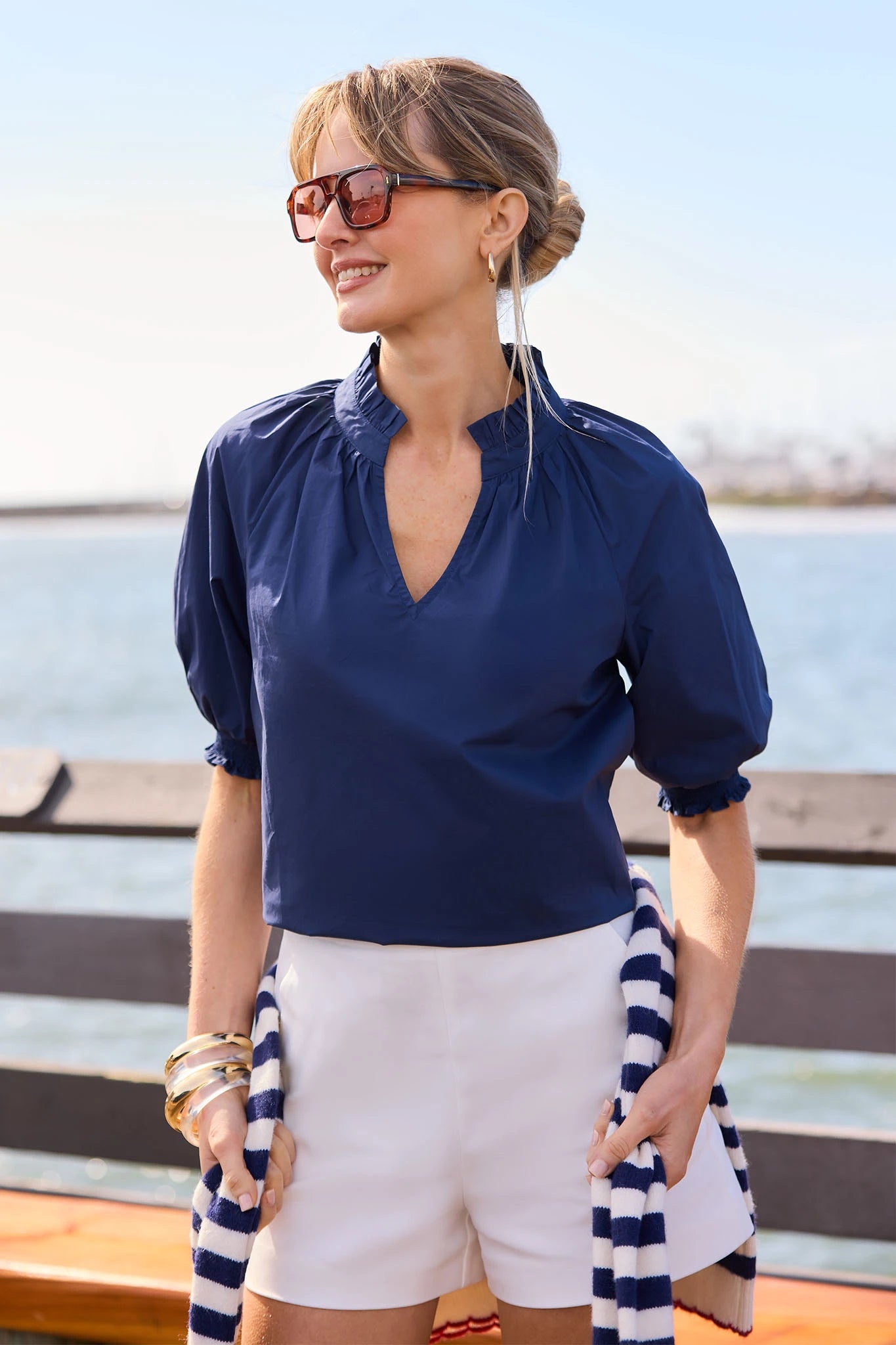 Woman wearing a navy blue top and white shorts with a striped cover-up by the water.