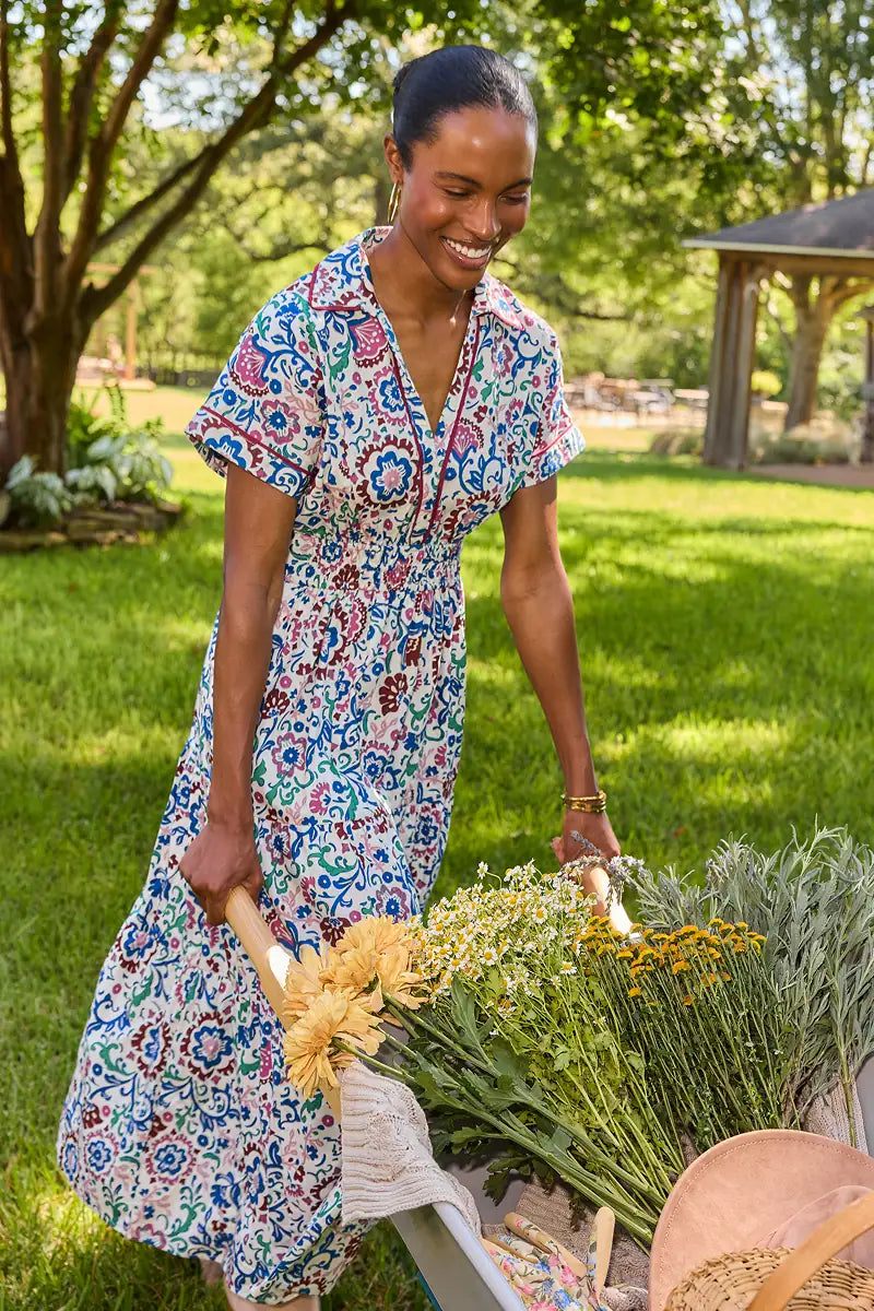 A woman in a short-sleeve V-neck maxi dress with a colorful paisley pattern pushes a wheelbarrow filled with dried flowers in a sunny, grassy garden.