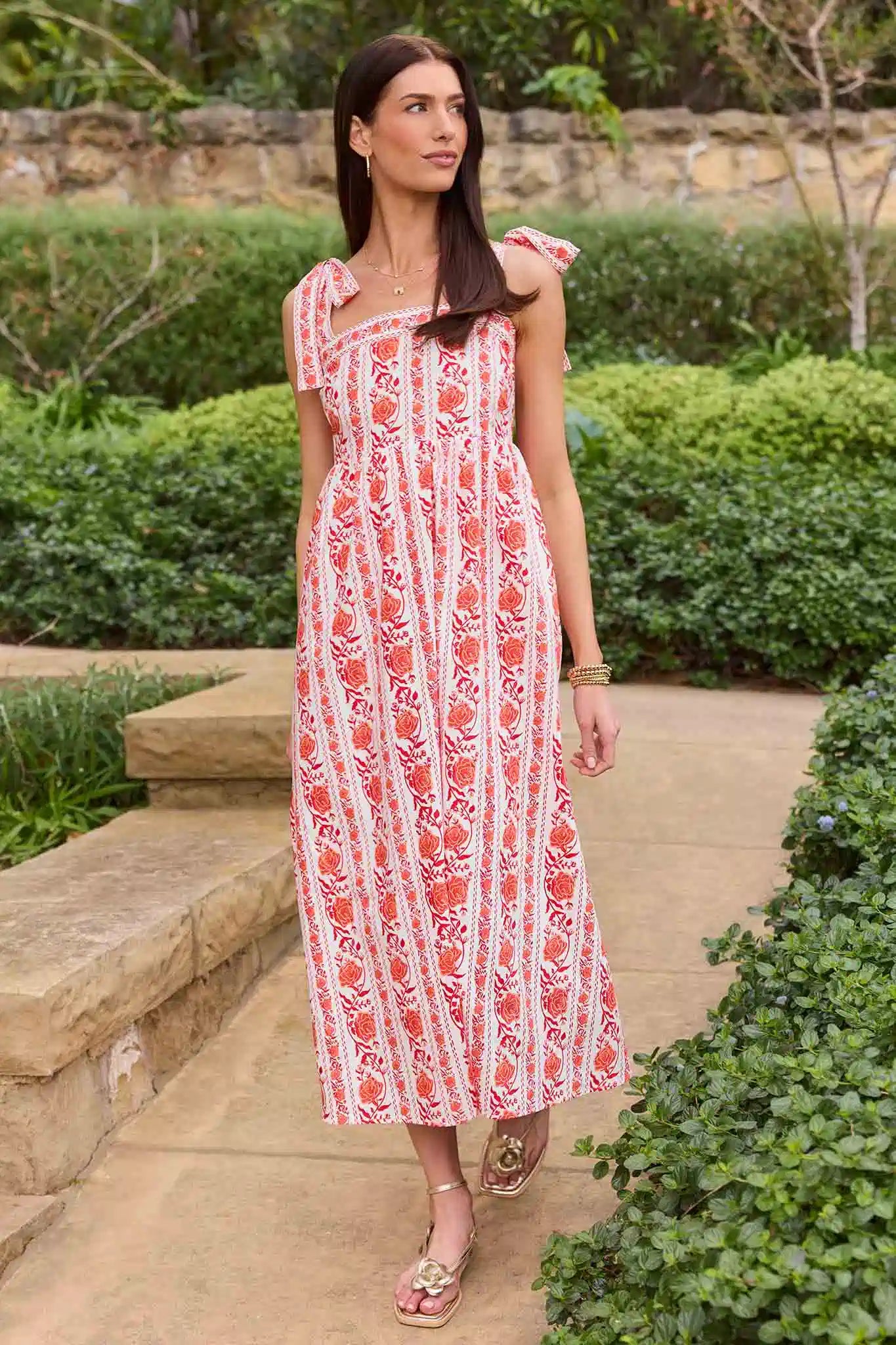 Woman in a red and white patterned dress standing outdoors with greenery around.