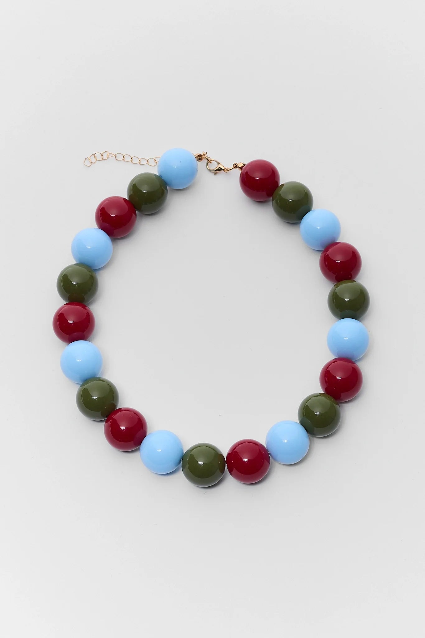A beaded necklace with alternating light blue, dark red, and olive green round beads, finished with a gold-tone clasp and extender chain, displayed on a light gray background.