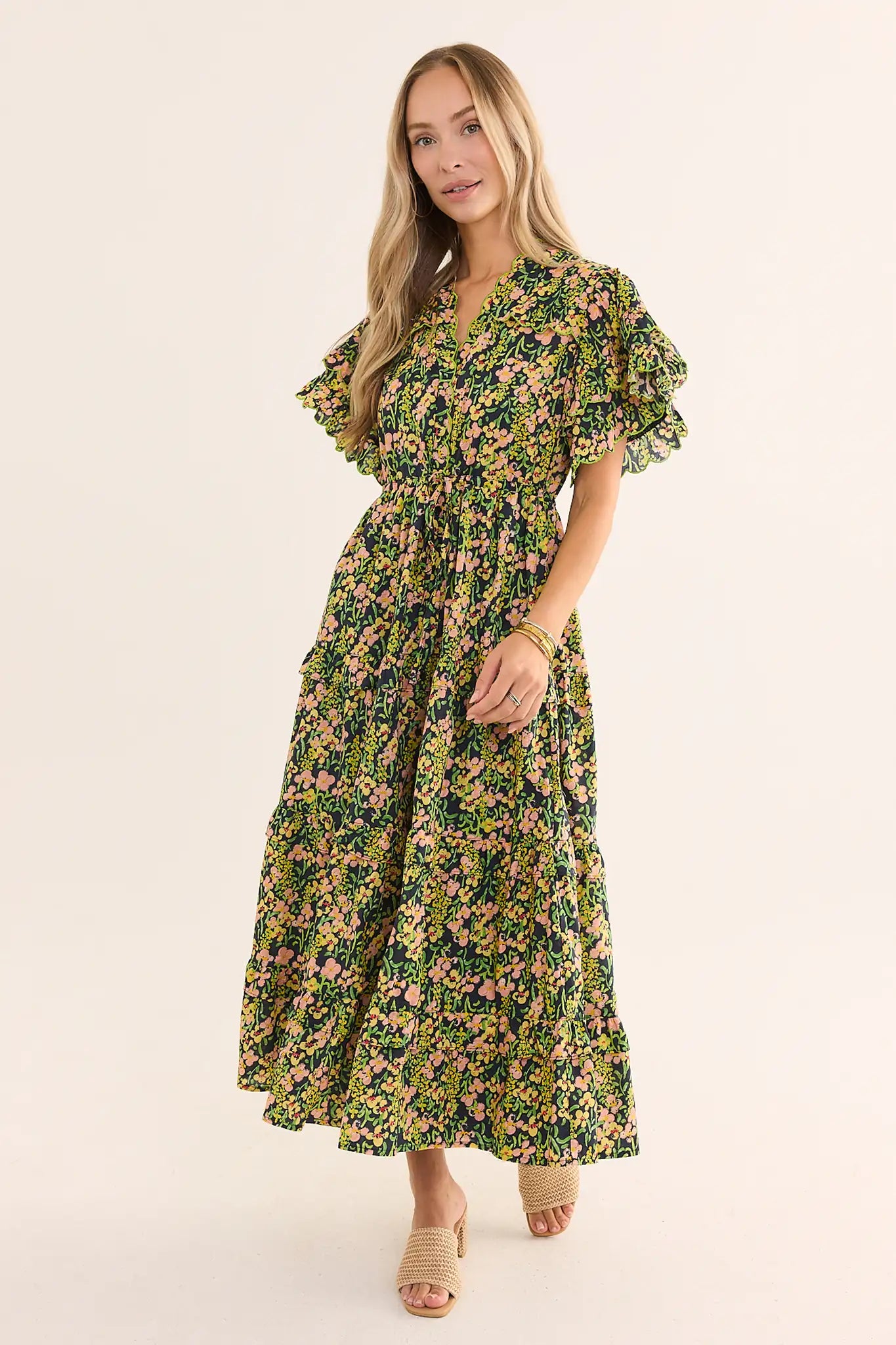 A woman modeling a long-sleeved, V-neck maxi dress with a green, yellow, and pink floral print, paired with beige slide sandals, against a plain light beige studio background.