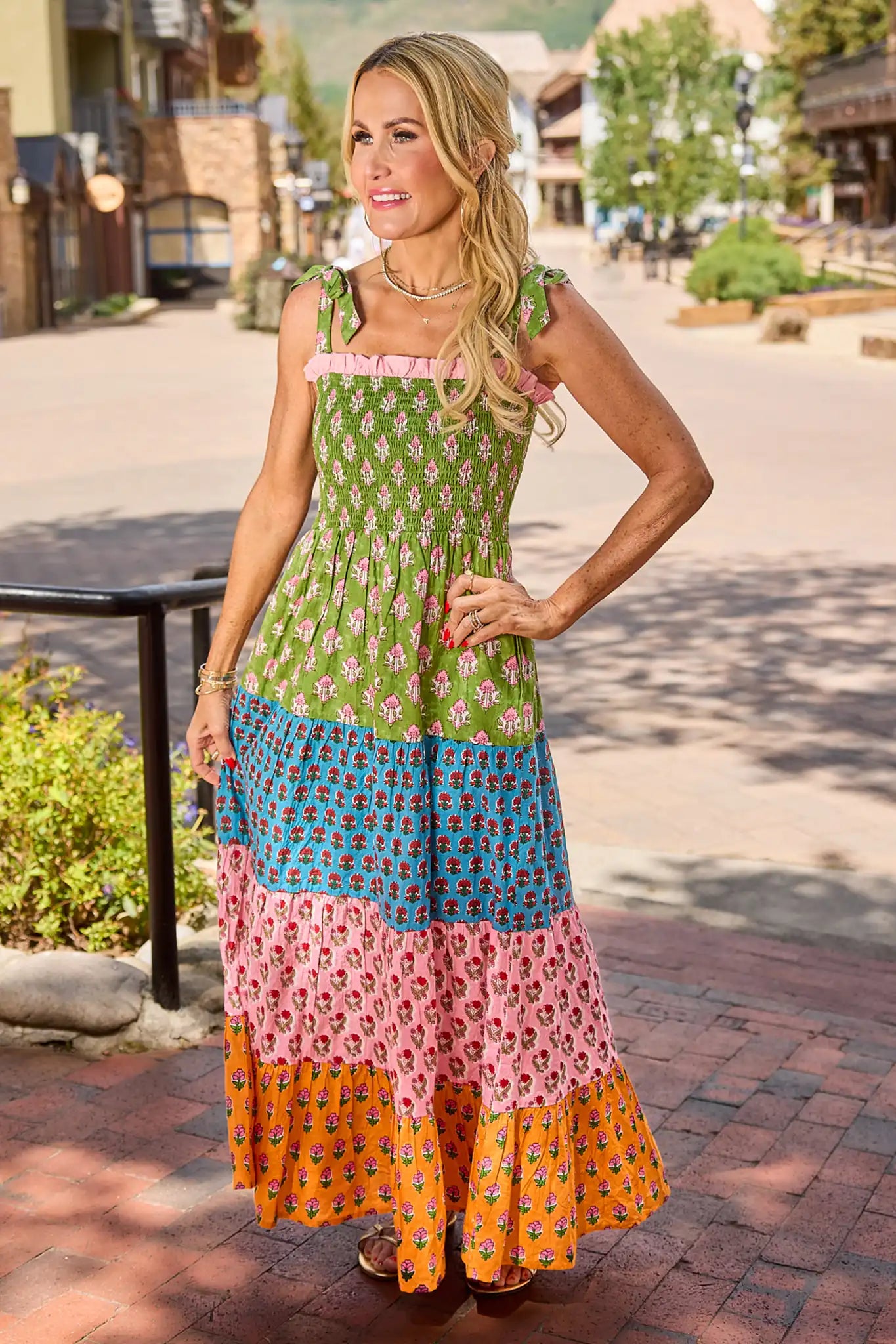 A woman modeling a sleeveless tiered maxi dress with a colorful patchwork print, standing on a brick walkway in an outdoor village setting.
