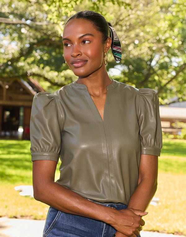 A woman wearing a short-sleeve olive green faux-leather blouse with puffed sleeves, standing outdoors in a sunny park setting.