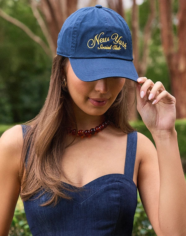 Woman wearing a blue cap with 'New York Social Club' text, standing outdoors.
