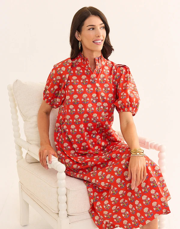 Woman wearing a red floral dress sitting on a white chair against a white background nav
