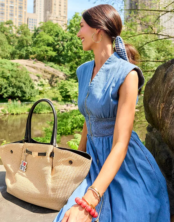 A woman in a blue denim dress sits outdoors near a park pond, next to a woven beige handbag with black handles and a small  charm. Trees, rocks, and city buildings are visible in the background. navigation