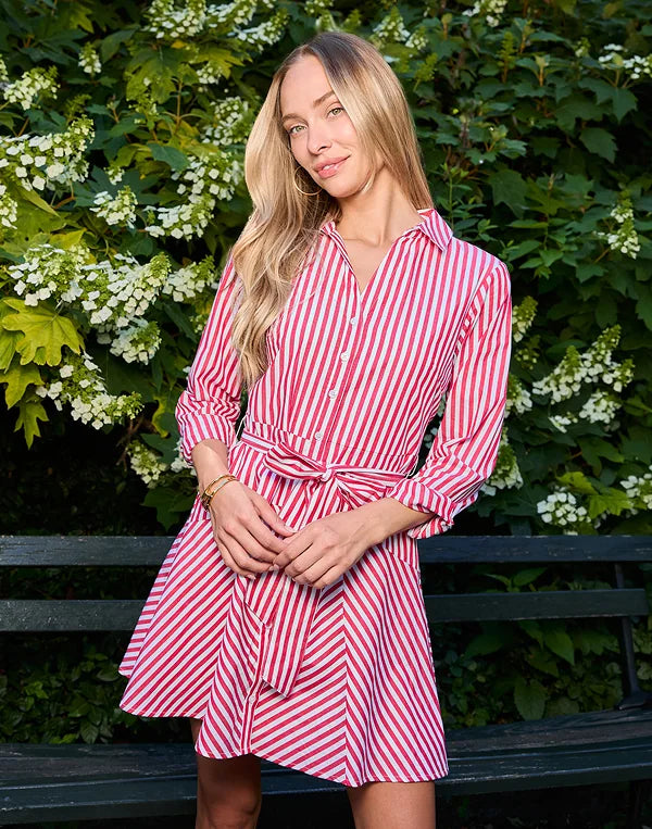 A woman wearing a red and white vertically striped shirt dress, standing outdoors in front of a green leafy hedge and a dark bench.