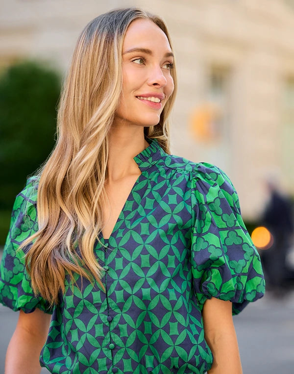 A woman with long blonde hair wearing a green and navy blue geometric pattern blouse with puff sleeves, photographed outdoors against a softly blurred background. navigation