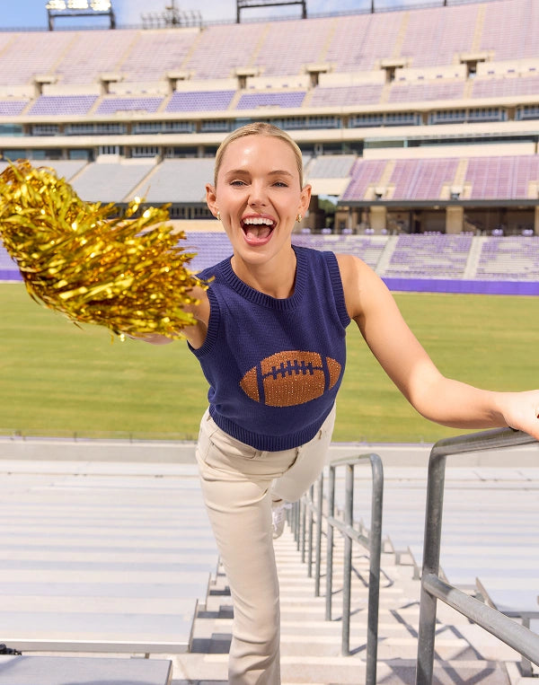 A cheerleader in a navy blue sleeveless sweater with a football graphic and beige pants, holding a gold tinsel pom-pom while standing on stadium bleachers in a large outdoor sports arena.