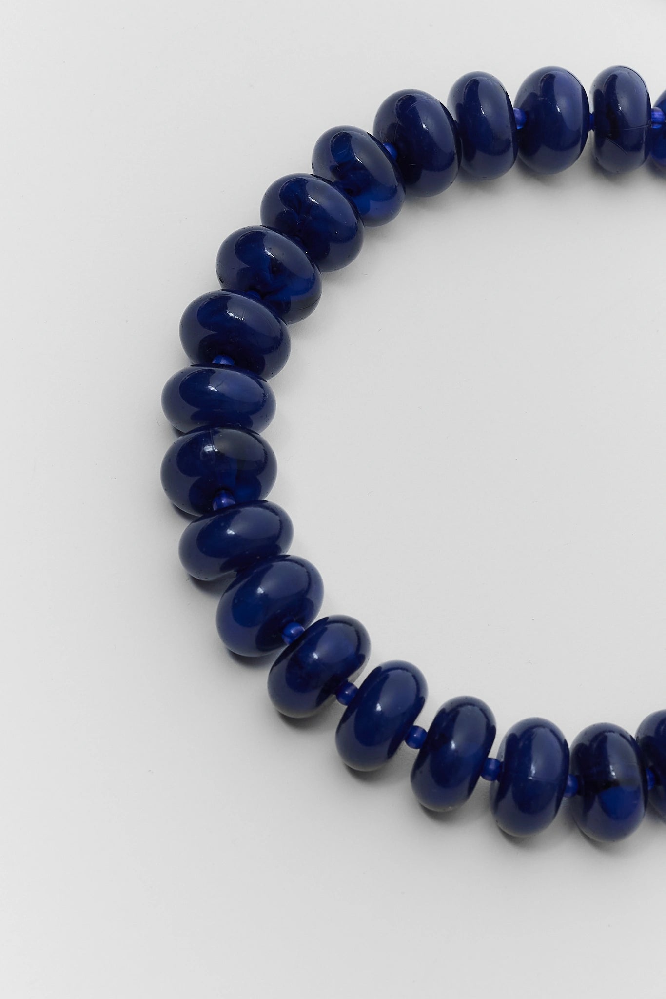 A close-up of a beaded necklace or bracelet made of glossy, dark blue oval beads on a plain light gray background.