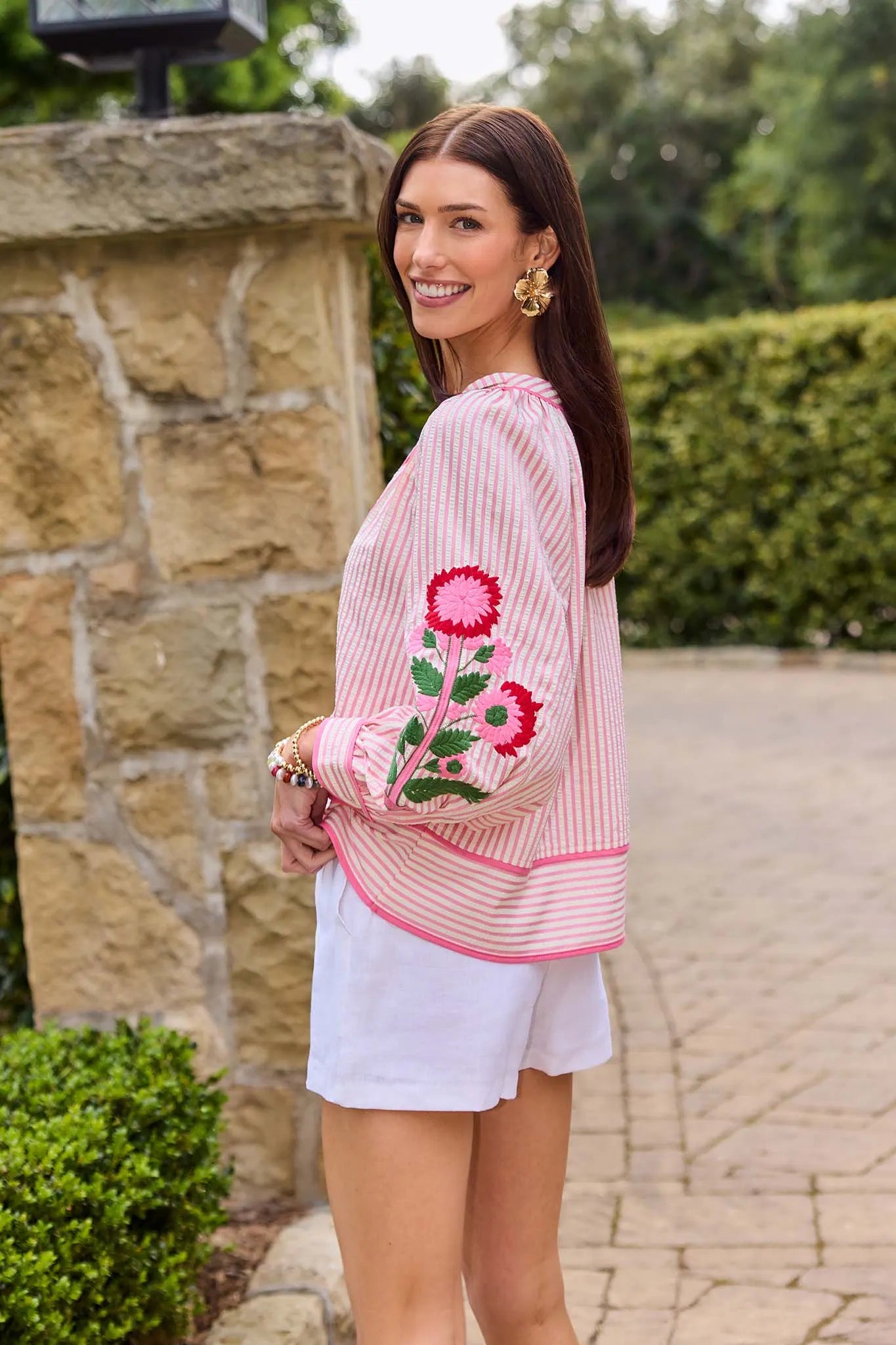 Woman wearing a pink embroidered top and white shorts standing outdoors.