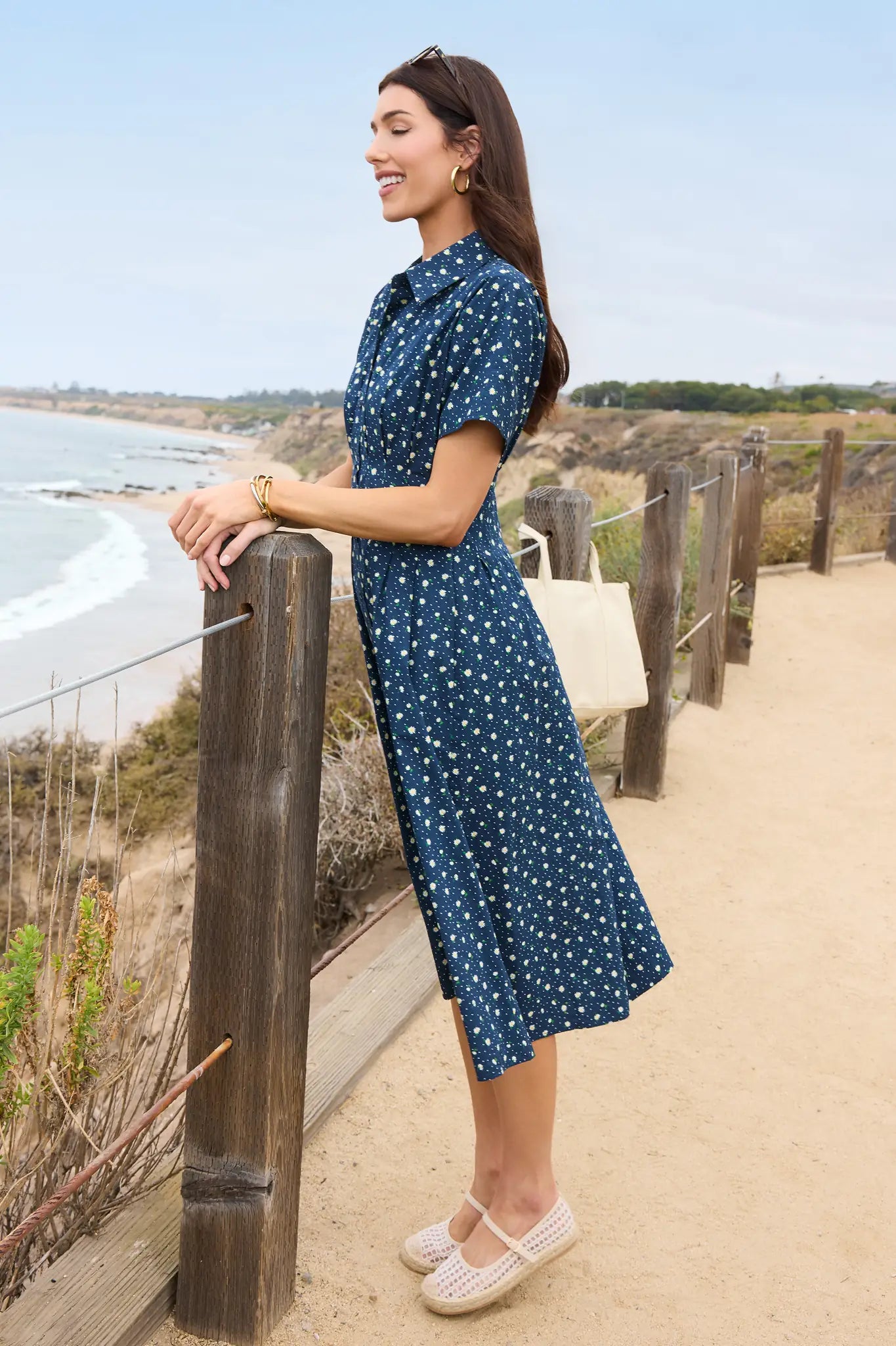 Woman in a blue polka dot dress standing by a wooden railing on a beach.