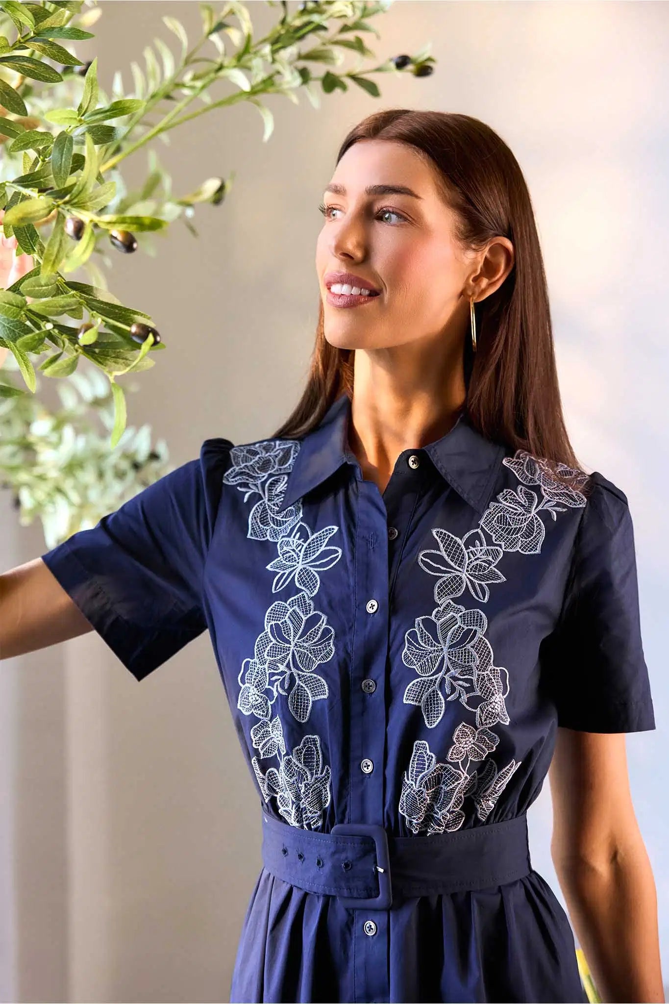 Woman wearing a navy blue dress with floral embroidery, standing indoors with plants in the background.